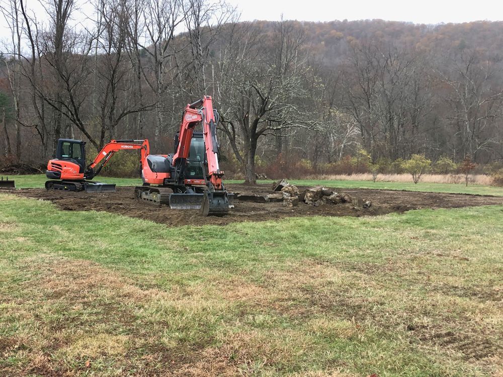Two orange excavators on a dirt patch in a grassy field. Trees and a hill in the background.