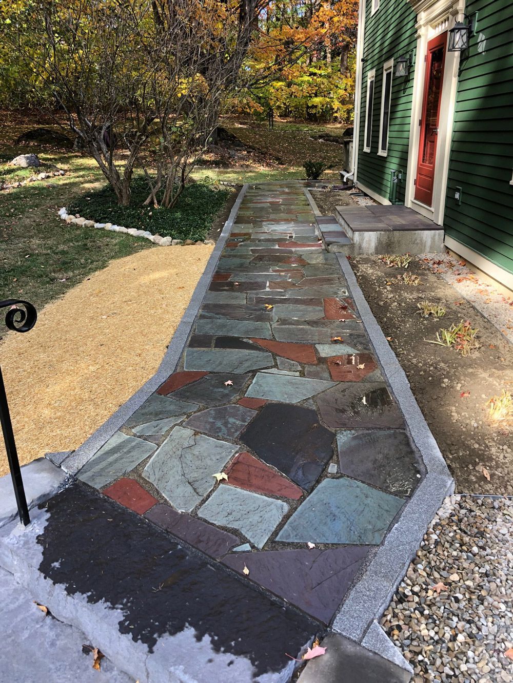Stone walkway leading to a green house with red door. Sunlight falls on the pavement.