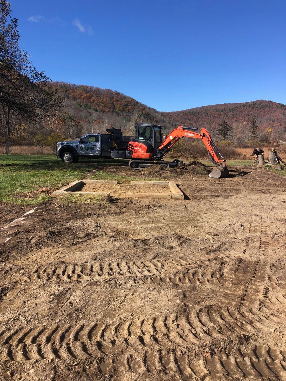 A small excavator digging at a construction site with a truck, under a blue sky, near a mountain.