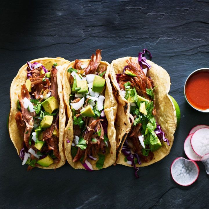 Three tacos with meat, avocado, cabbage, and radishes on a table.