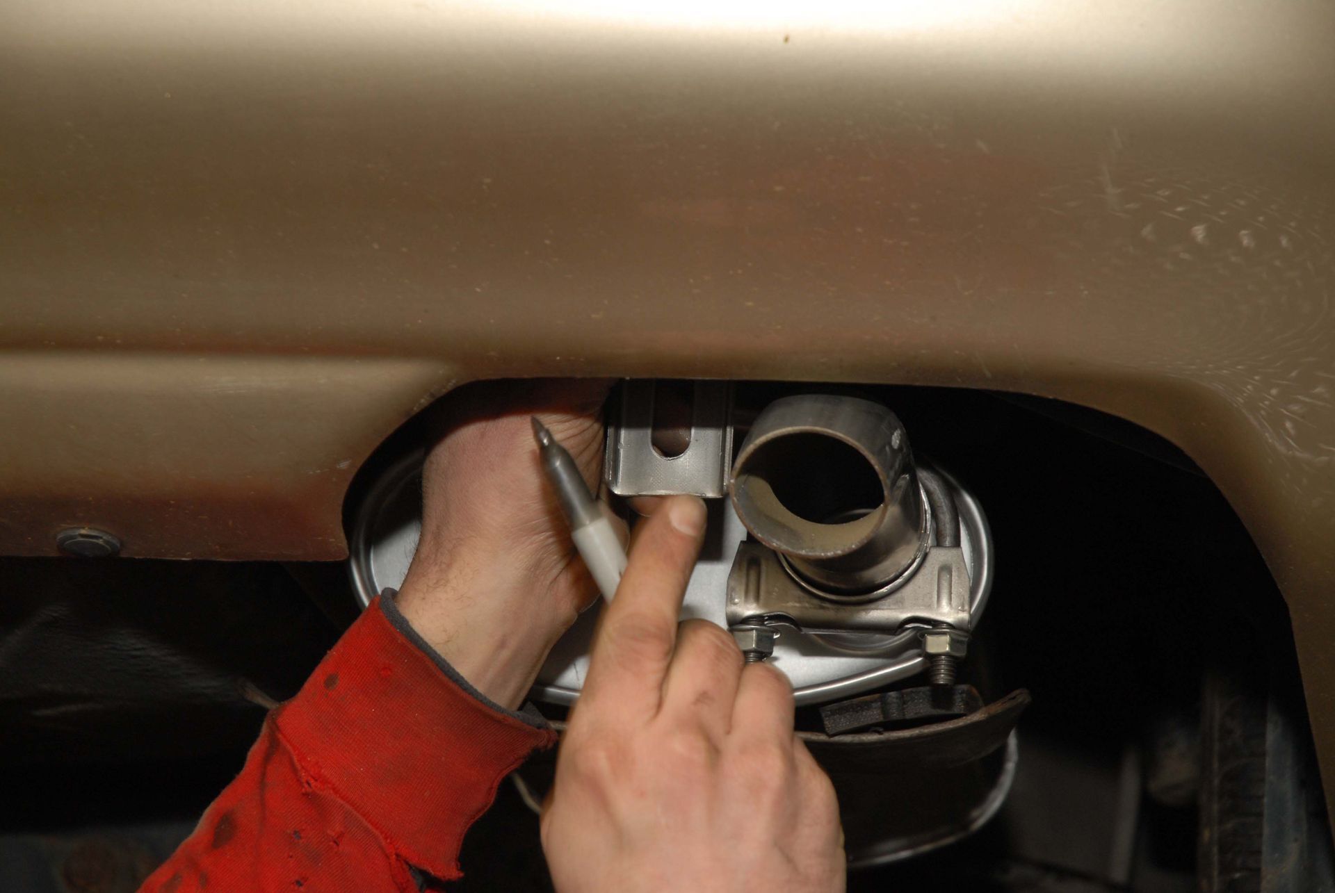 Person using a pen to mark a metal bracket connected to a car's exhaust system.
