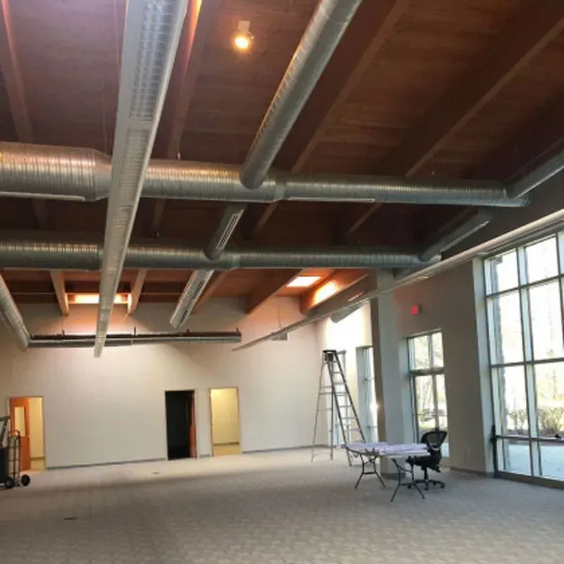 Interior of a room with exposed ductwork on a wood ceiling and large windows. Empty space with a ladder.