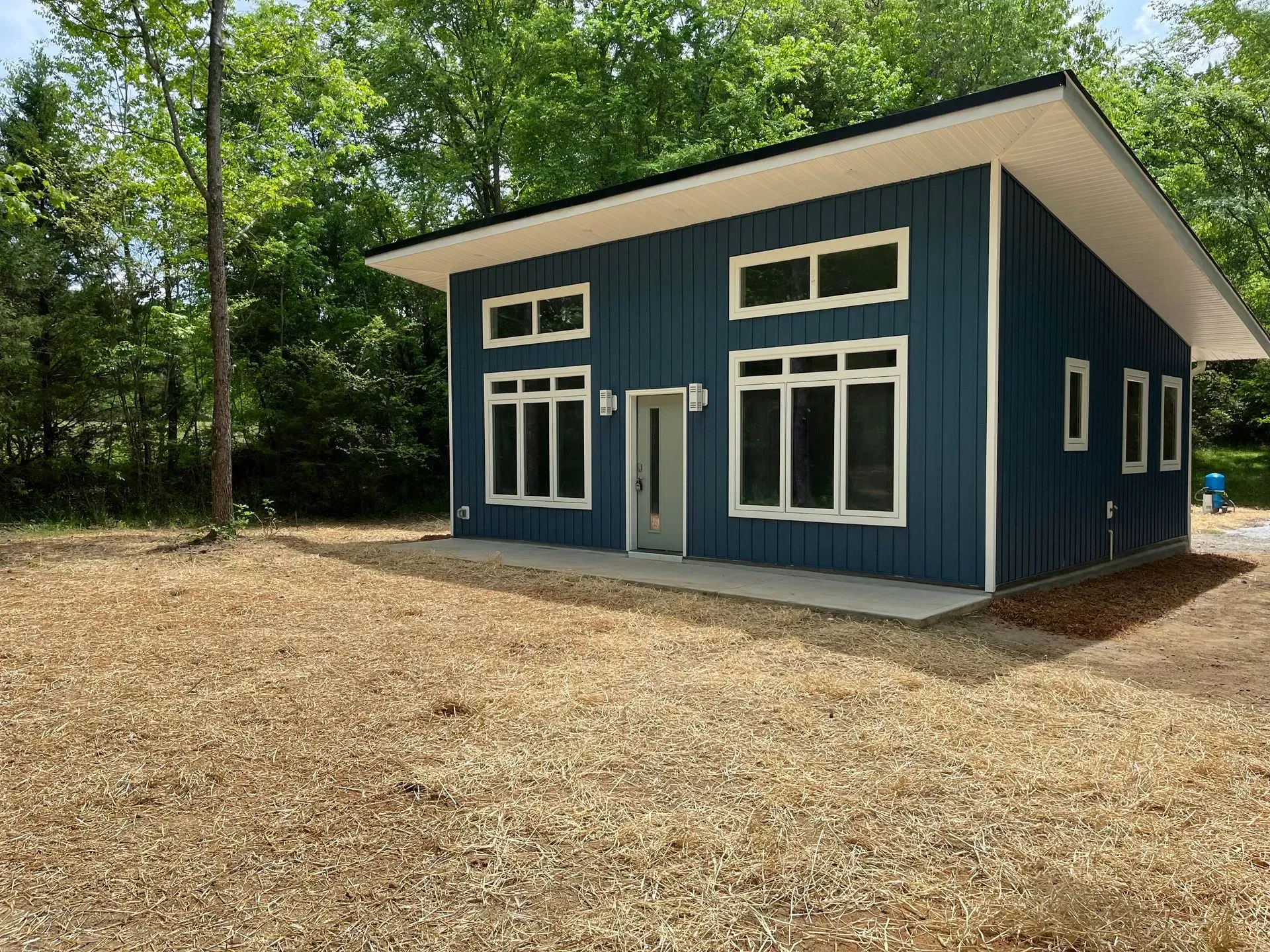 Blue modern cabin with white trim and large windows, surrounded by gravel and trees.