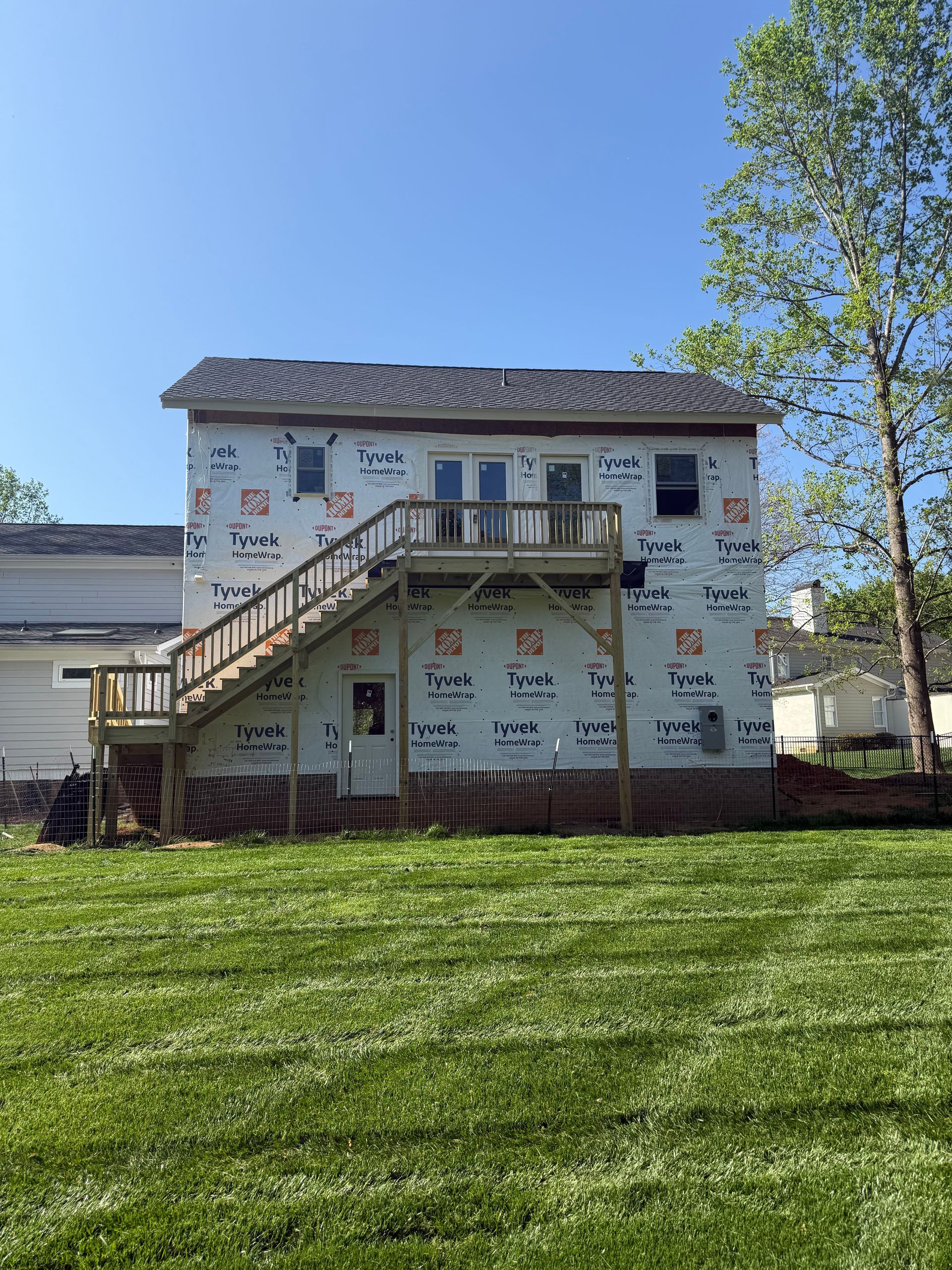 A two-story house under renovation, covered in white vapor barrier sheeting with a wooden outdoor staircase and deck.
