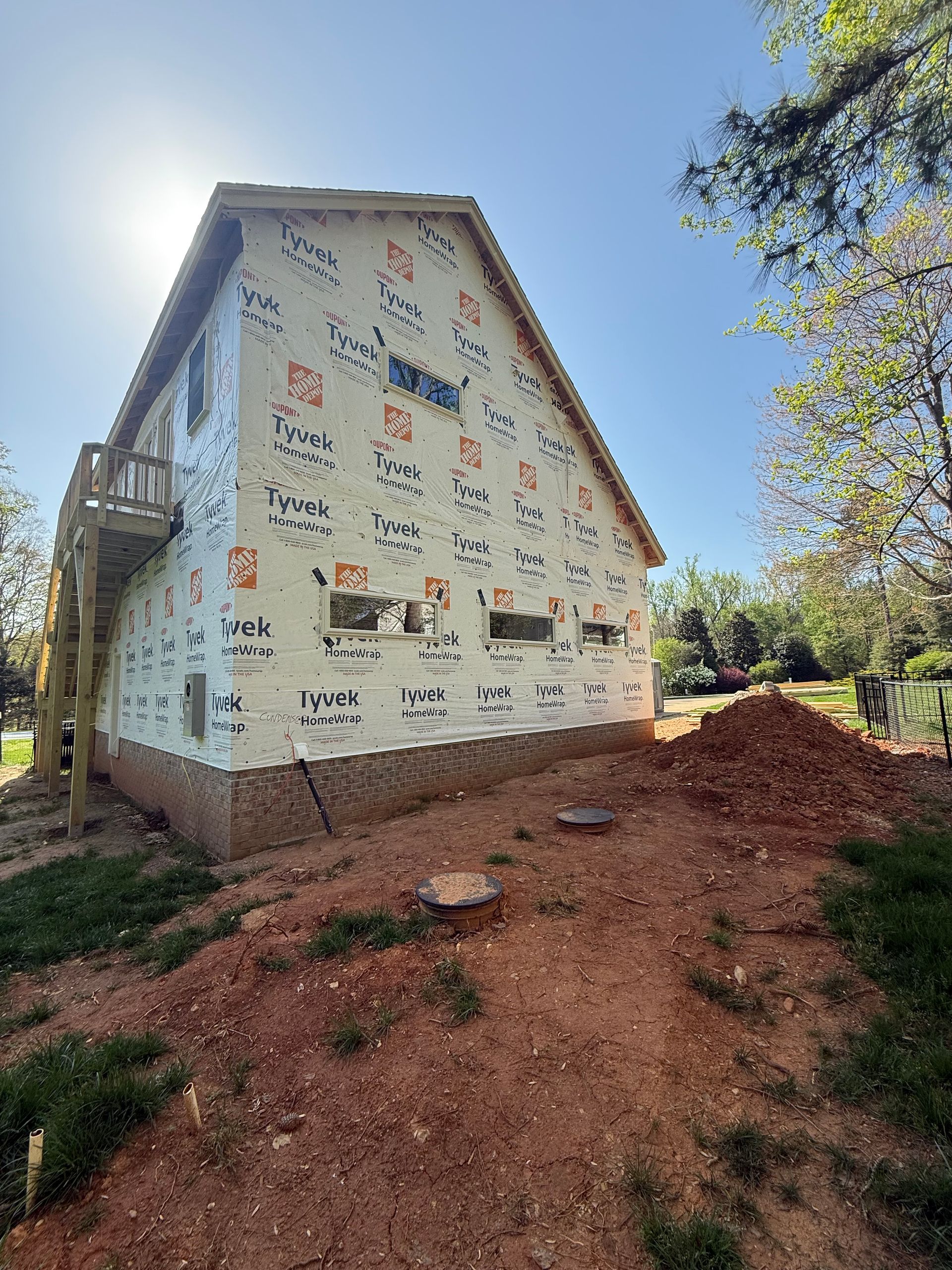 A side view of a house under construction, covered in white Tyvek wrap, with a brick foundation and a pile of dirt outside.