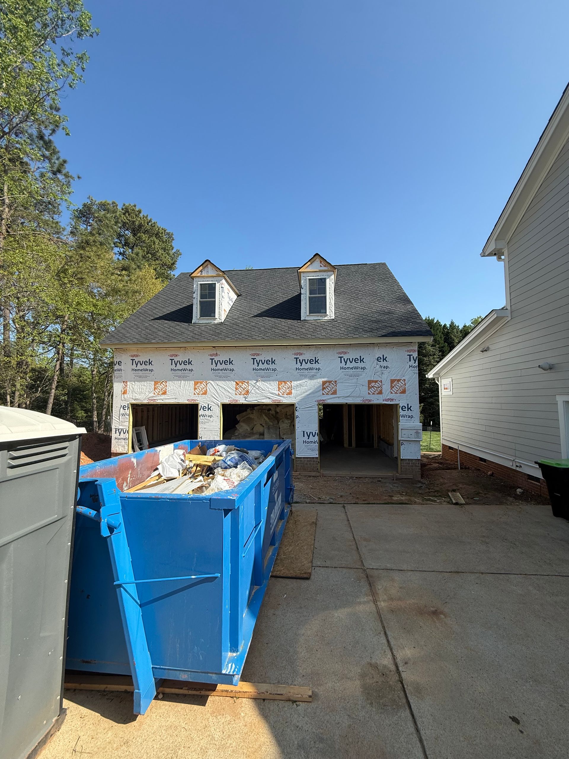 A blue dumpster sits in a driveway in front of a house under construction with exposed sheathing and dormer windows.