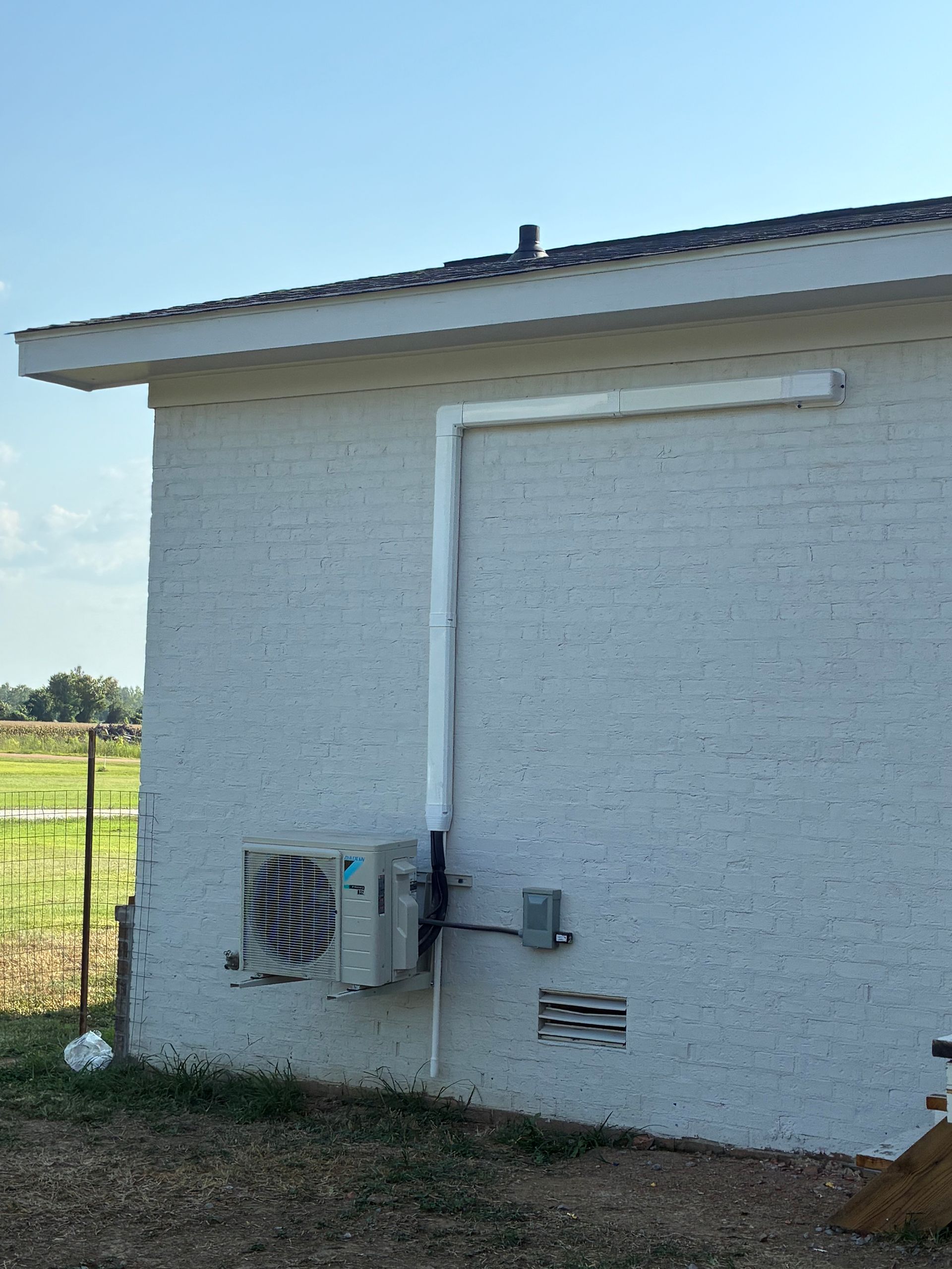 White brick building side with outdoor AC unit, white pipe, and vent under a sunny blue sky