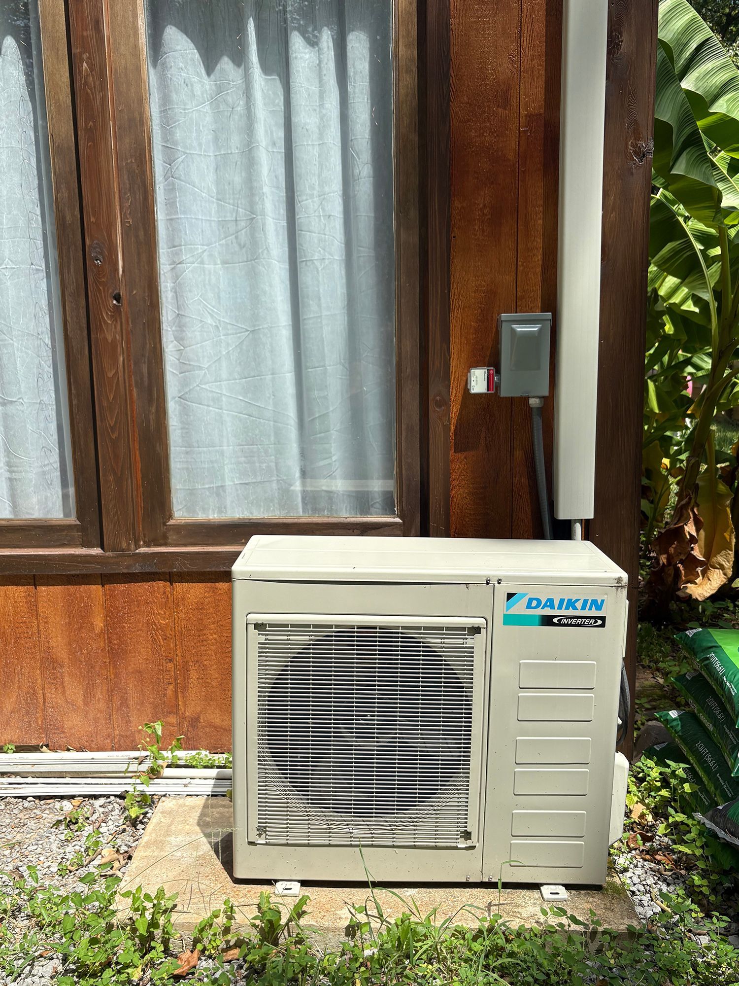 Outdoor air conditioner unit beside a wooden house wall and window curtains