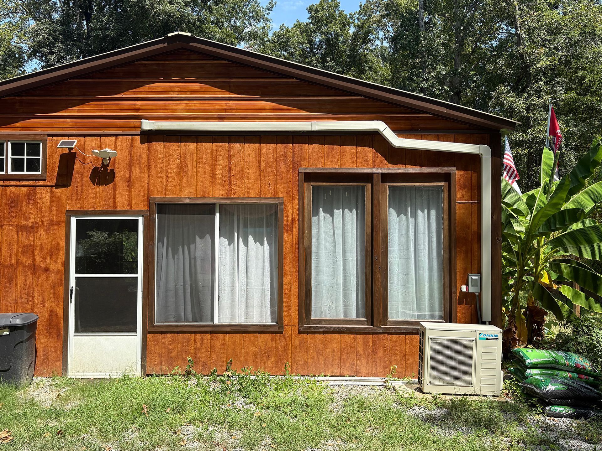 Wooden house exterior with white door, windows, and an air conditioner in a sunny yard