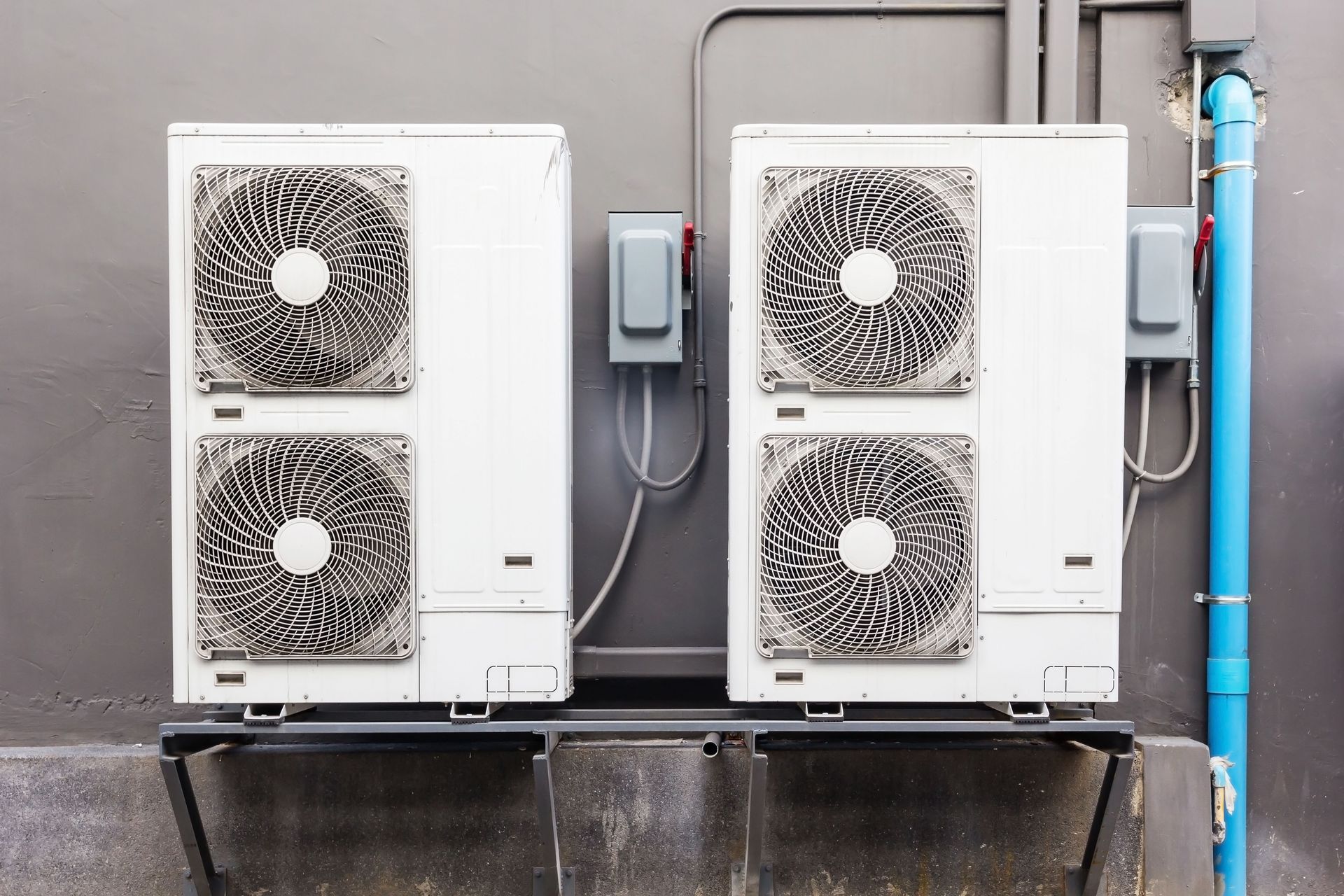 Two white outdoor HVAC units with fans, mounted against a gray wall next to blue piping.