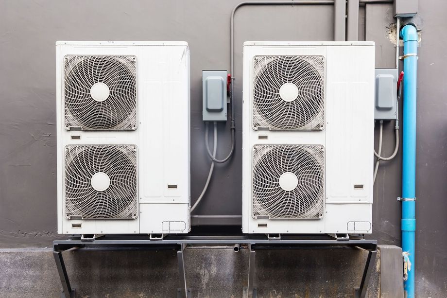 Two outdoor HVAC units mounted against a gray wall with pipes and cables nearby