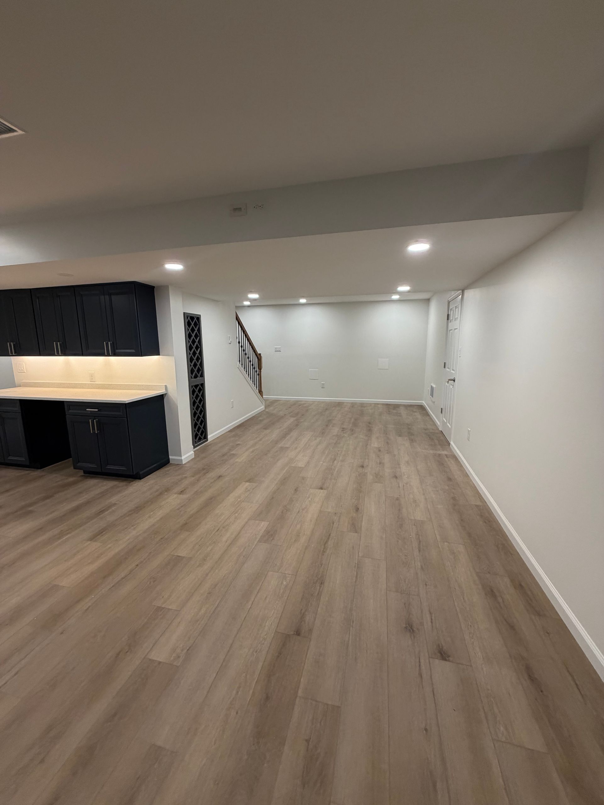 Empty basement room with wood flooring, white walls, recessed lights, and a dark built-in area on the left