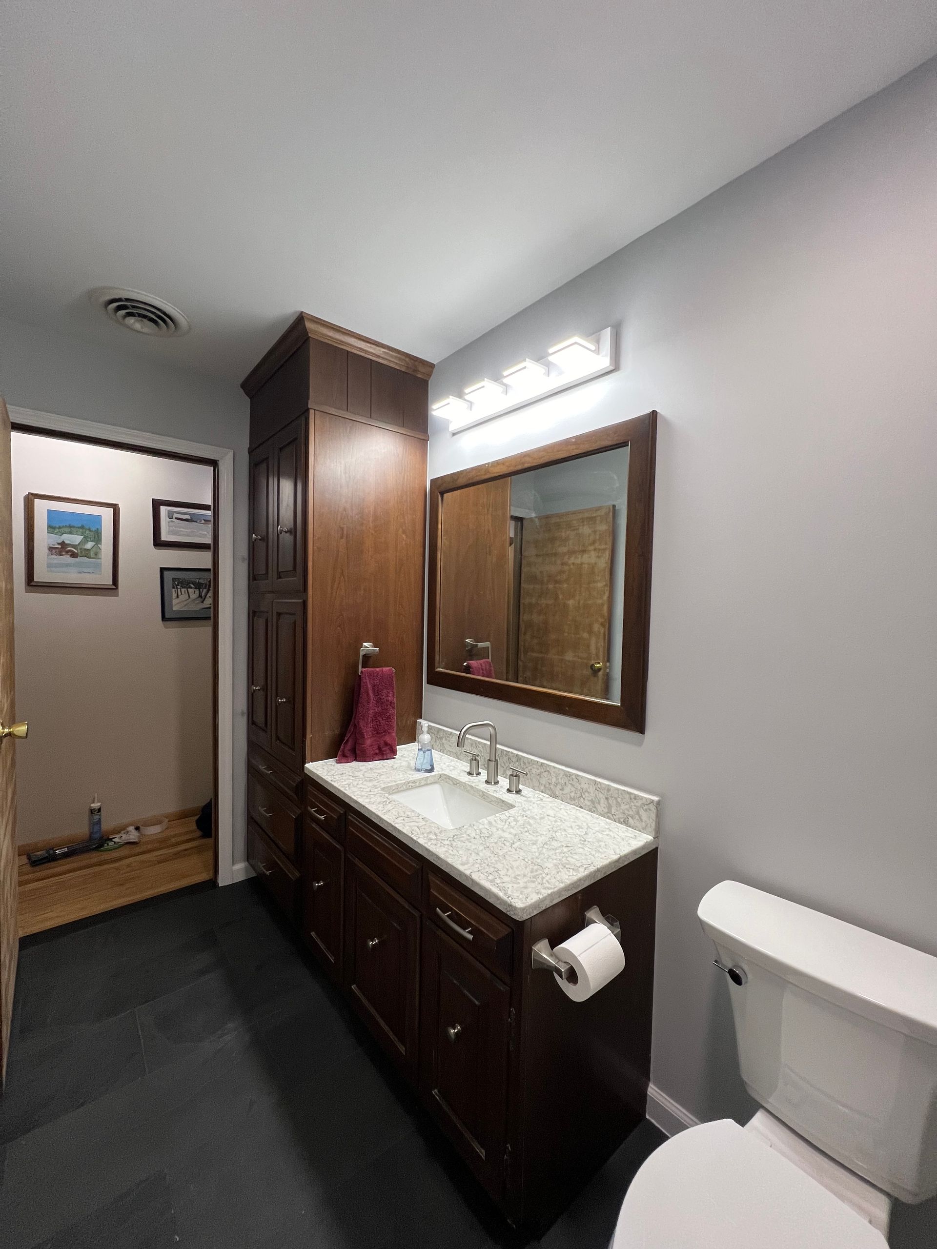 Bathroom with a dark wooden vanity, mirror, and toilet. Gray walls, dark floor, and a tall storage cabinet.