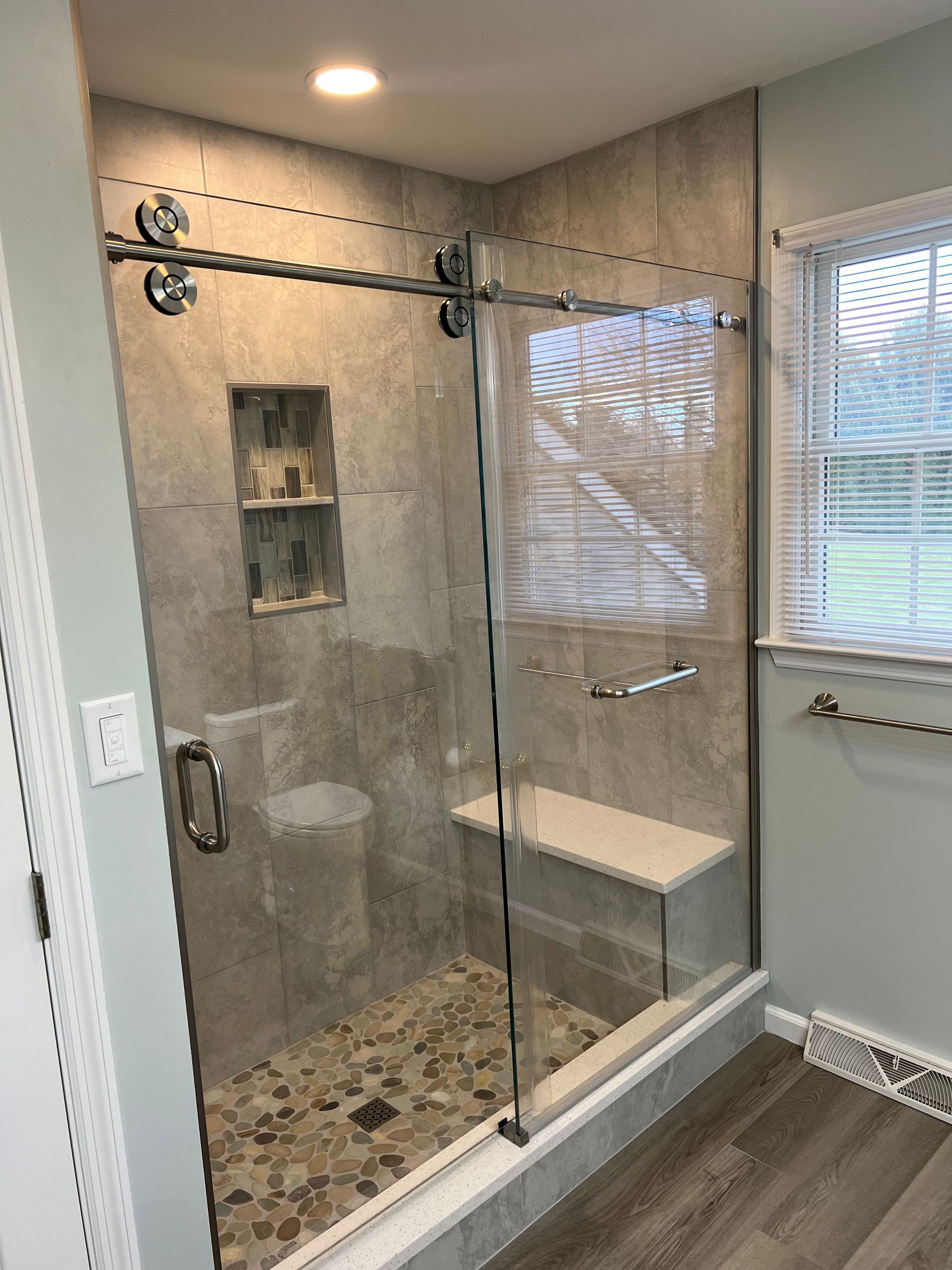 Modern walk-in shower with gray stone tile, glass door, and pebble floor. Features a built-in bench and recessed shelving.