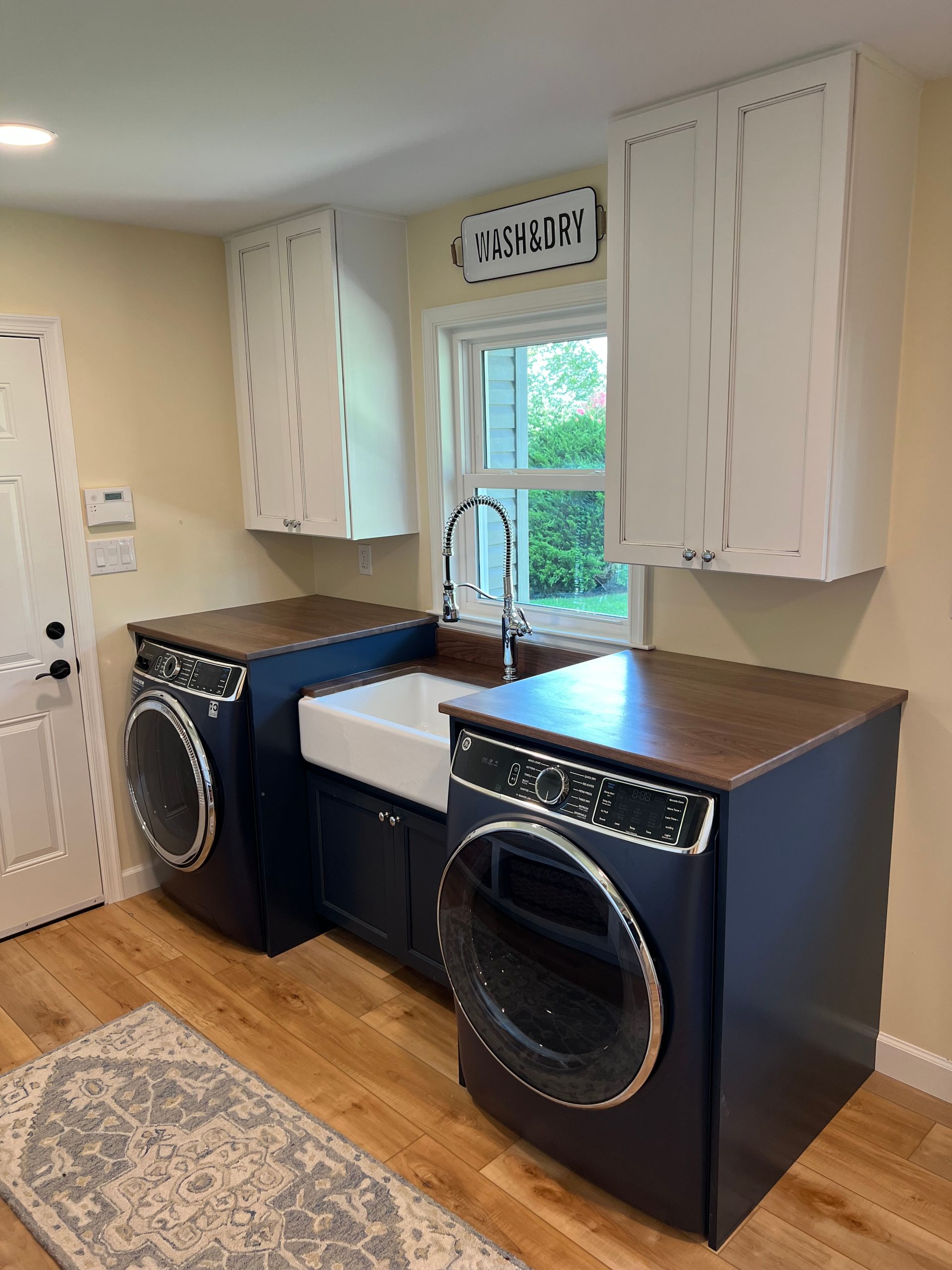 Laundry room with blue cabinets, white farmhouse sink, and washer/dryer units. Wooden countertop and white upper cabinets.