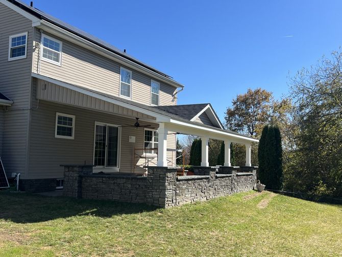 Two-story beige house with a stone-walled porch and a covered patio, under a clear blue sky.