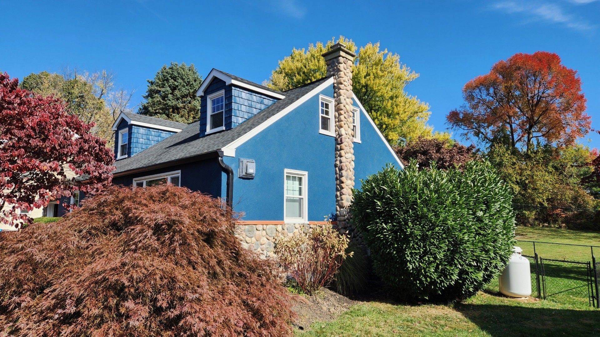 Blue house with dormers, brick chimney, and colorful fall foliage against a blue sky.