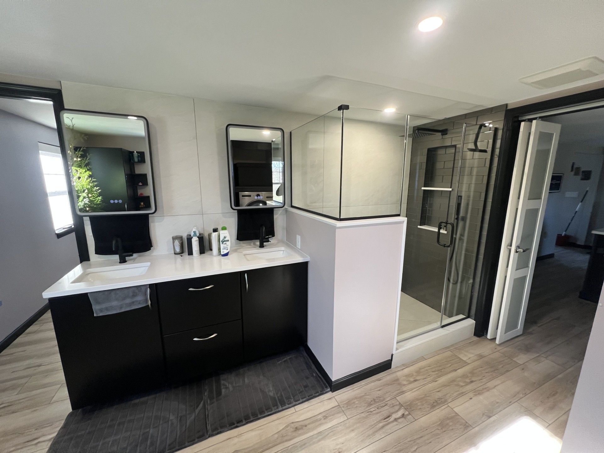 Bathroom with black vanity, white countertop, two mirrors, glass shower, and wood-look flooring.