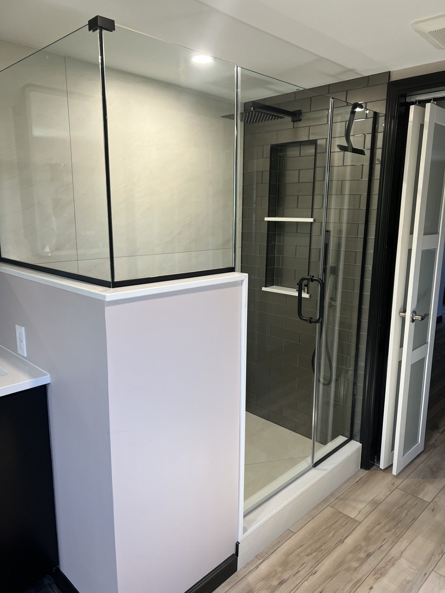 Modern shower with glass doors, black hardware, gray tile, and a white base on light wood flooring.