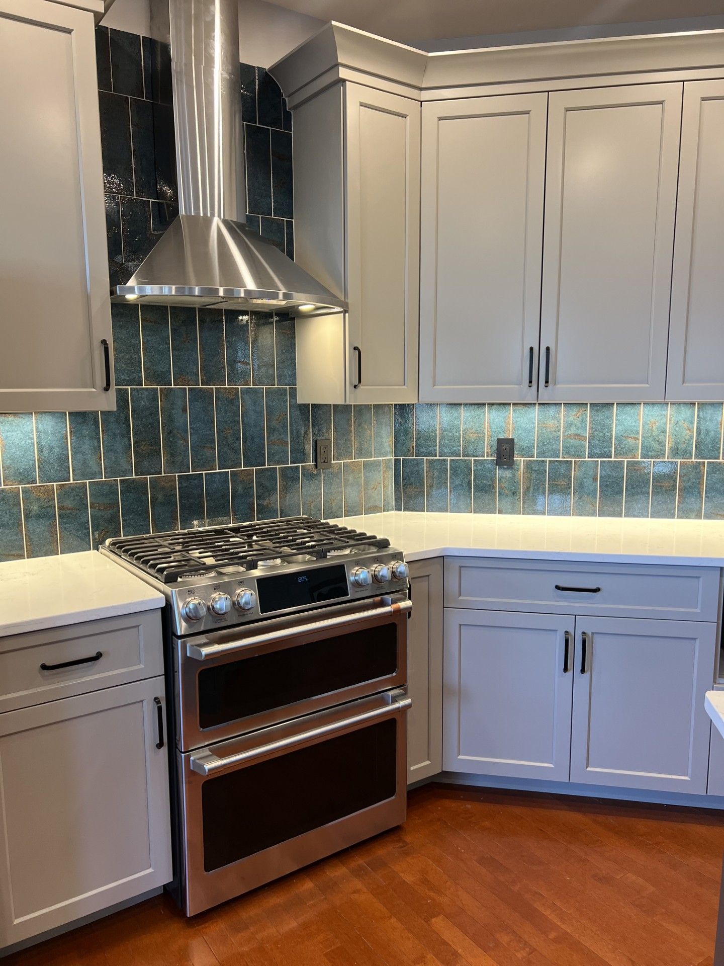 Kitchen with light gray cabinets, stainless steel range and hood, teal tile backsplash, and wood floors.
