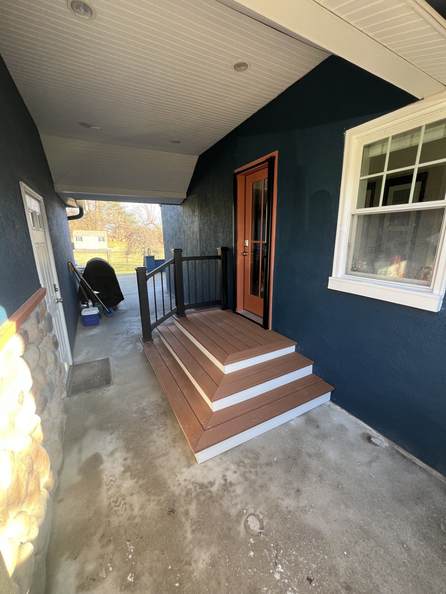 Exterior view with three composite steps leading to a wooden door. Dark blue siding, white trim, and a black metal railing are visible.