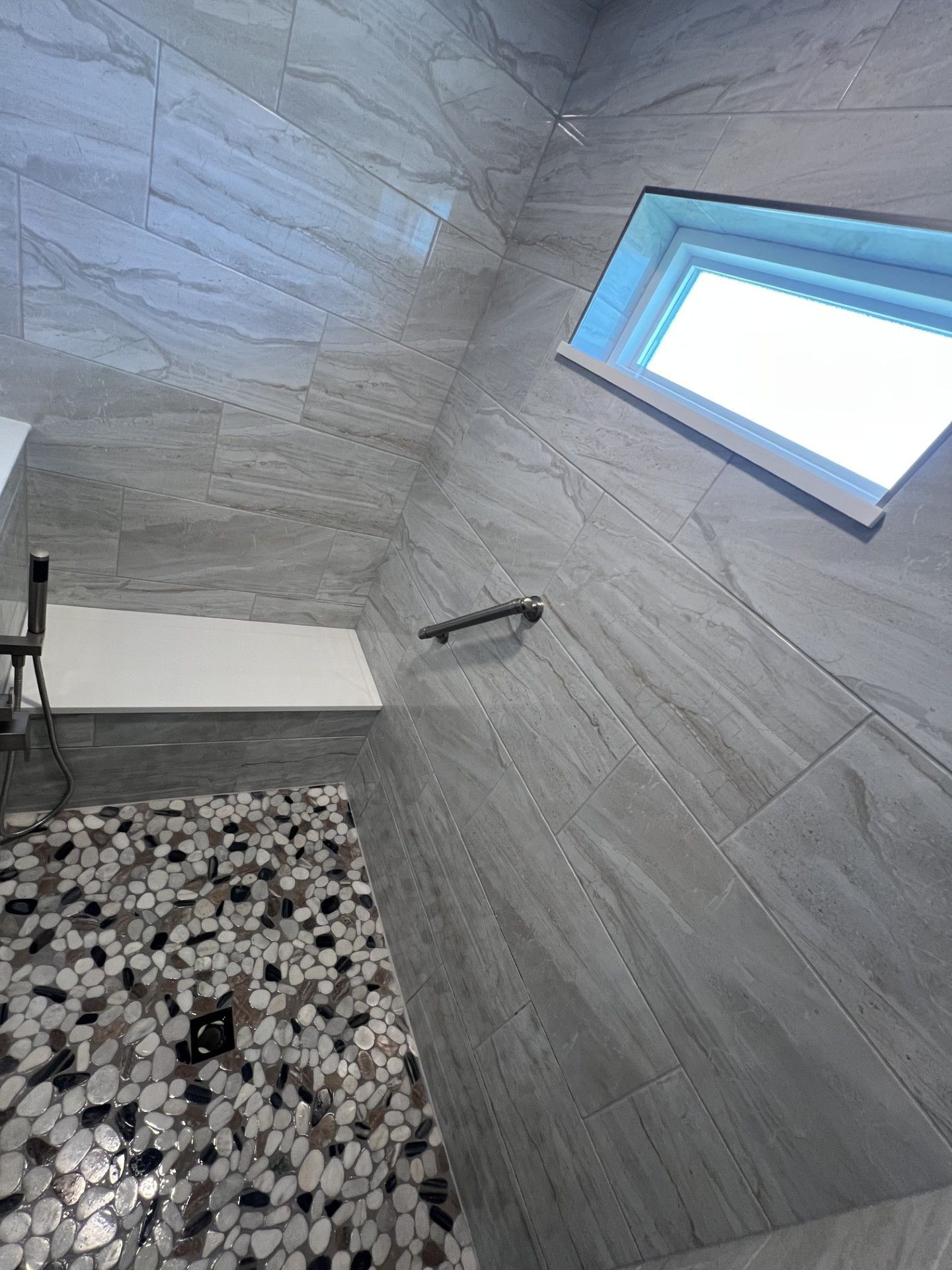 Shower with gray tiled walls, pebble floor, and window.