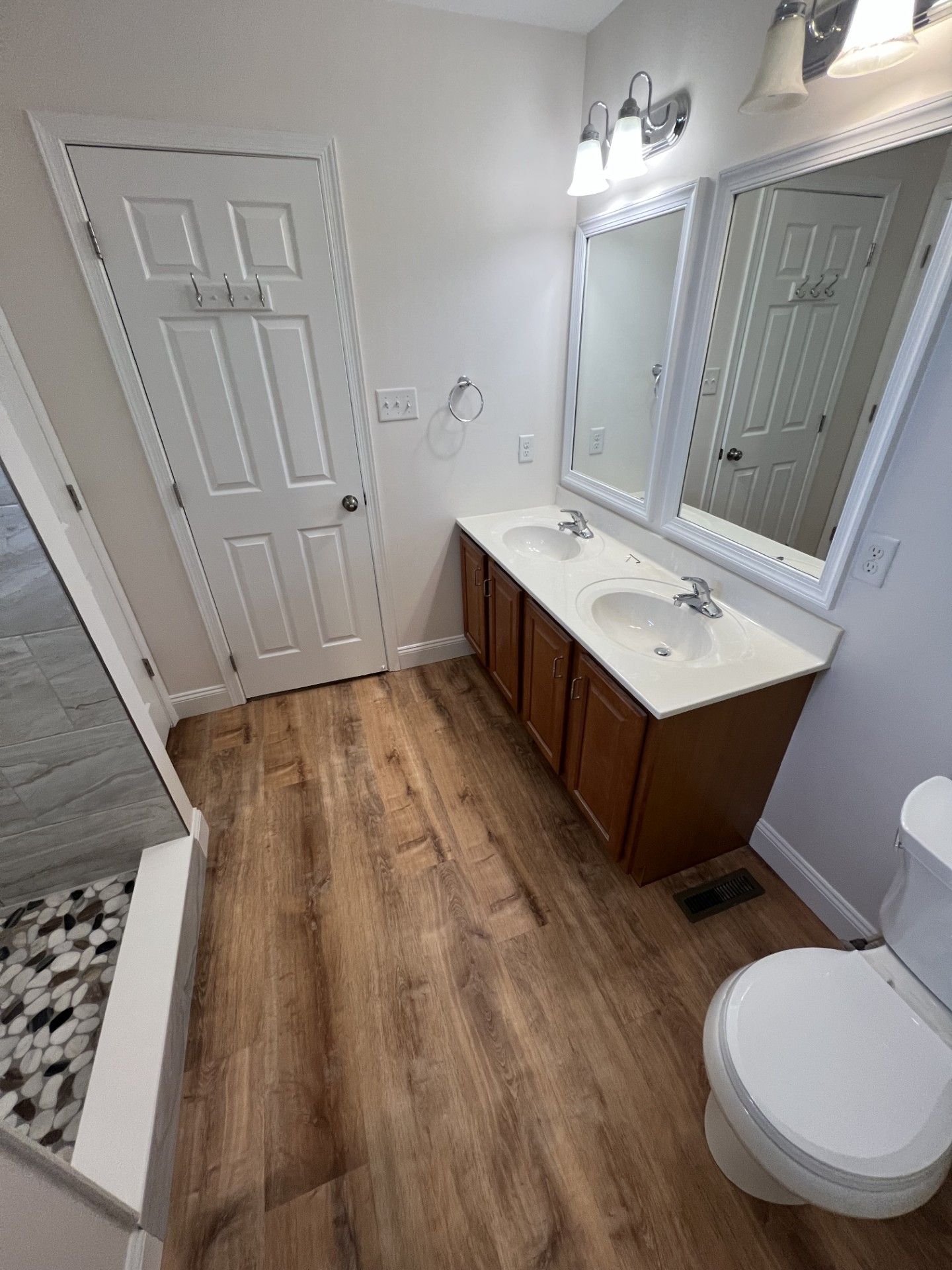 Bathroom with wood-look flooring, double sink vanity, toilet, door, and shower with pebble tile.
