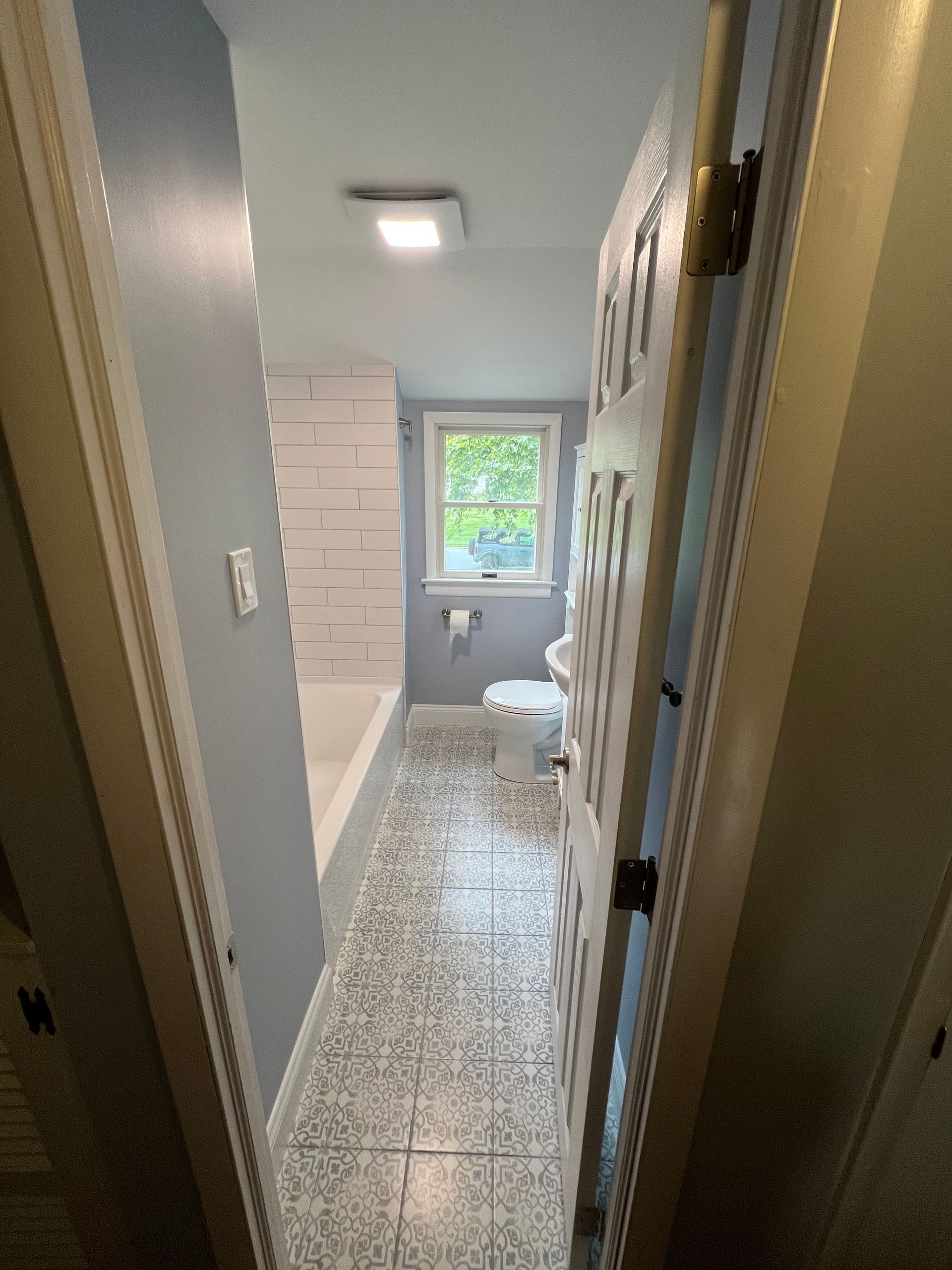 Narrow bathroom with light blue walls, white tub, patterned tile, and a partly open white door.