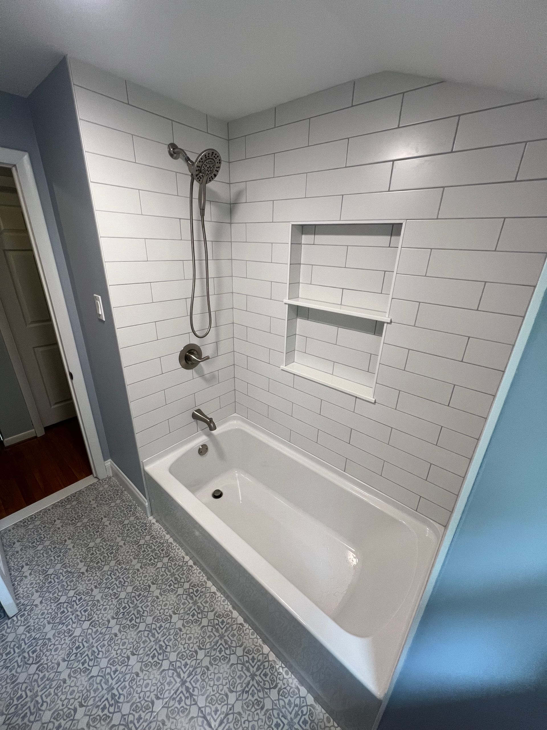 White-tiled bathroom with a bathtub, shower, and built-in shelf. Light blue walls, gray patterned floor.