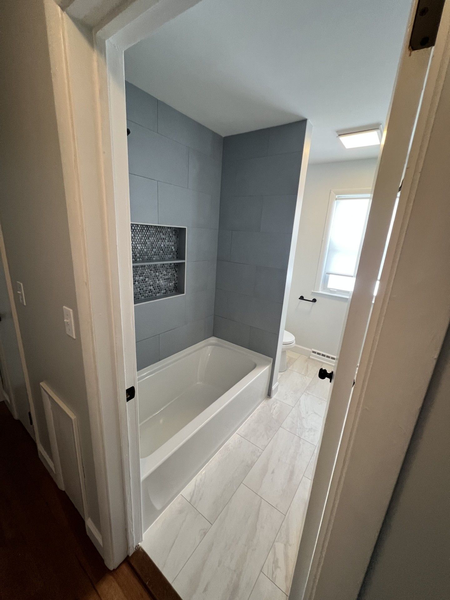 Bathroom with gray tiled shower and white tub, looking down a hallway with wood floor.