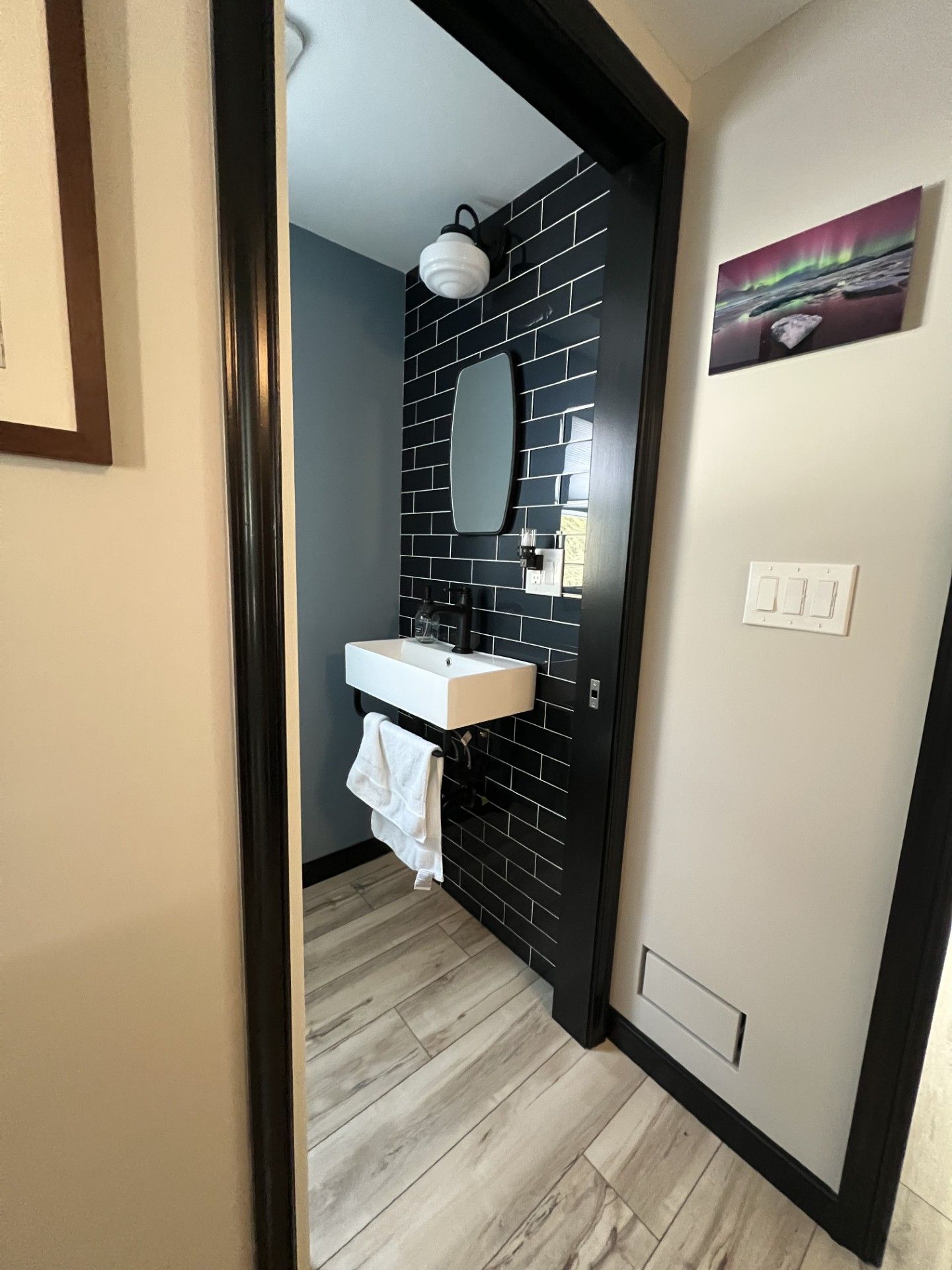 A small bathroom with black tile wall, white sink, oval mirror, and a wood-look floor.