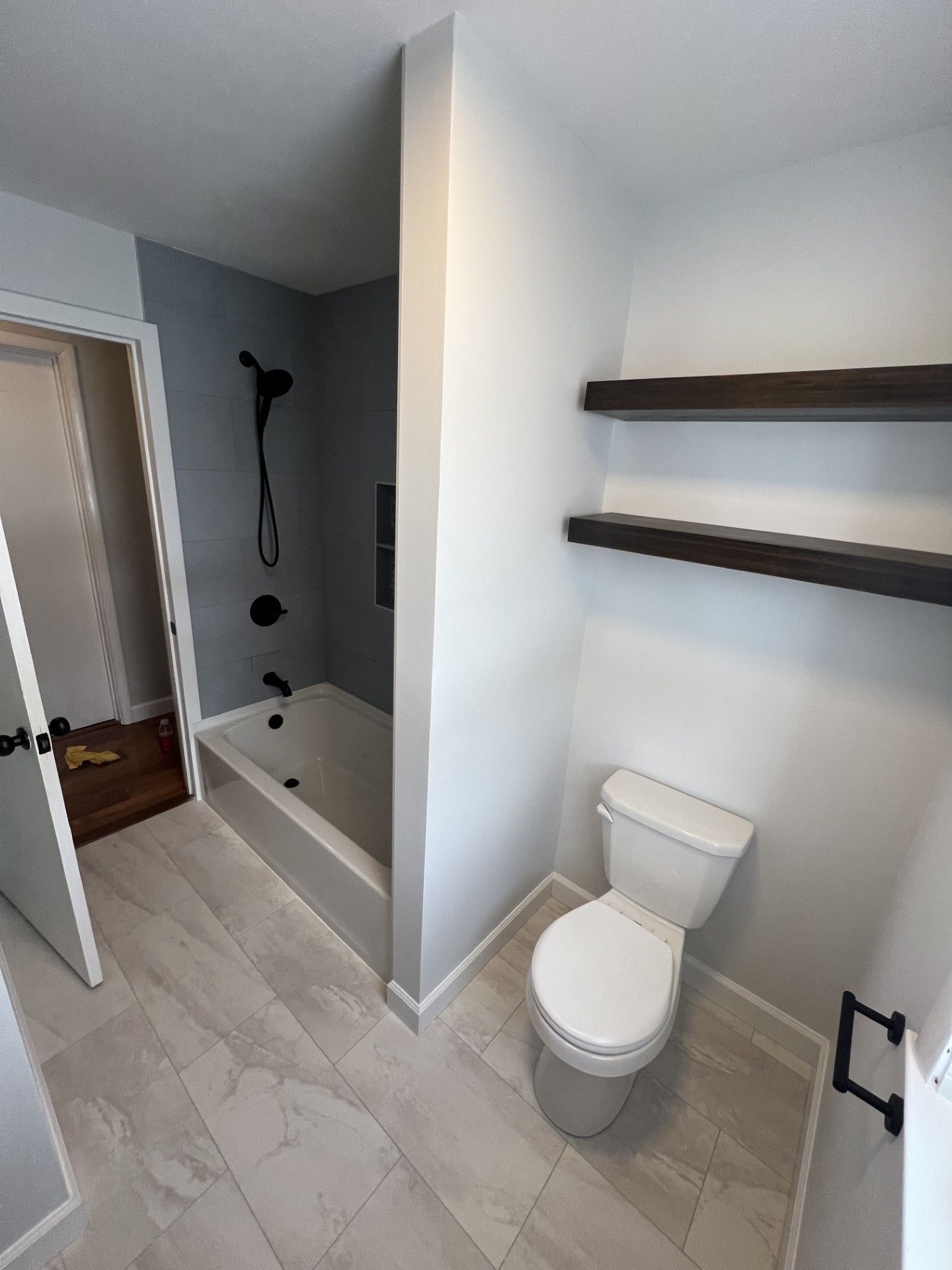 Bathroom with white walls, gray tile floor, toilet, dark shelves, and shower area.