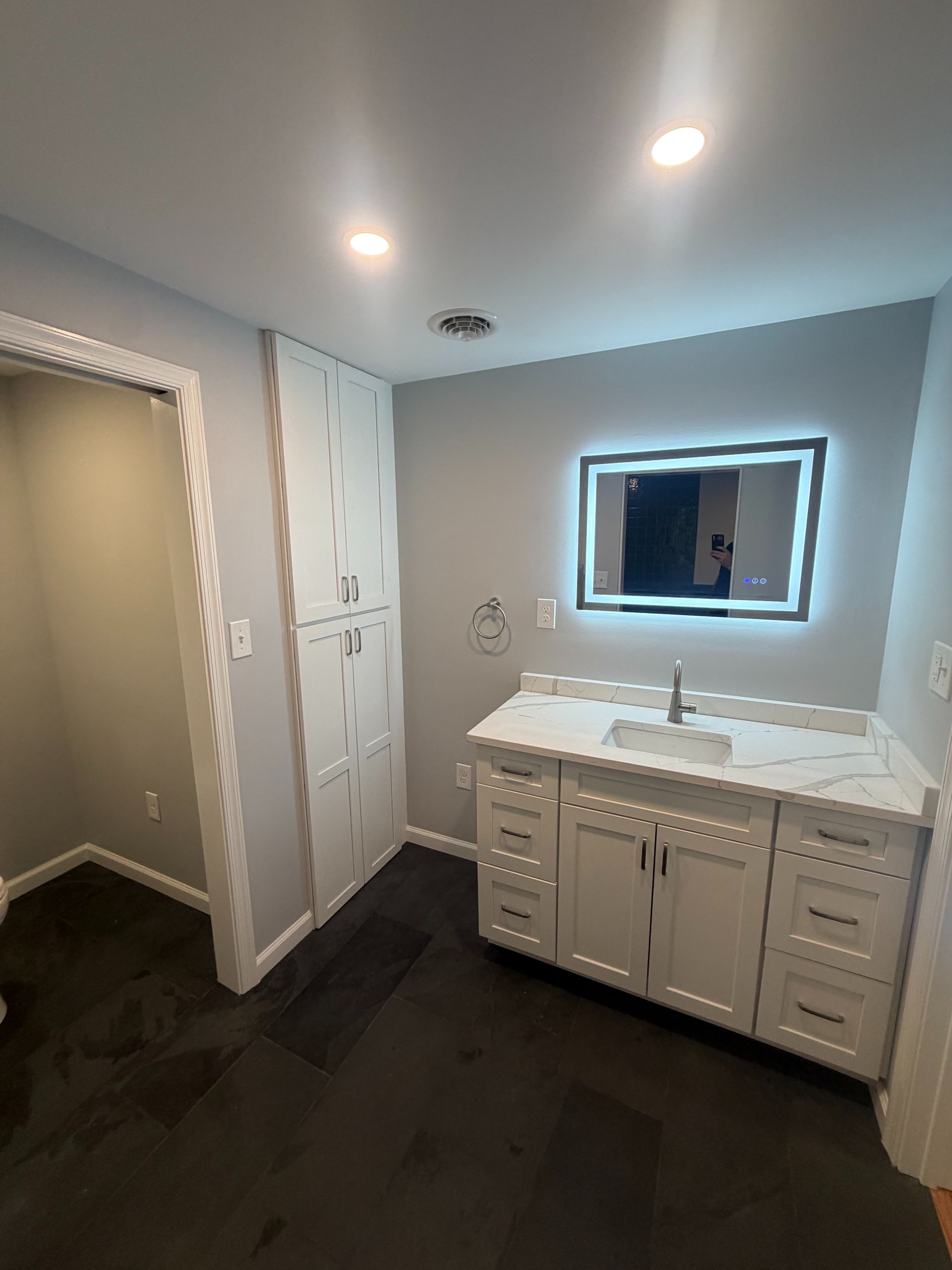 Modern bathroom with double vanity, illuminated mirror, white cabinets, and dark flooring.