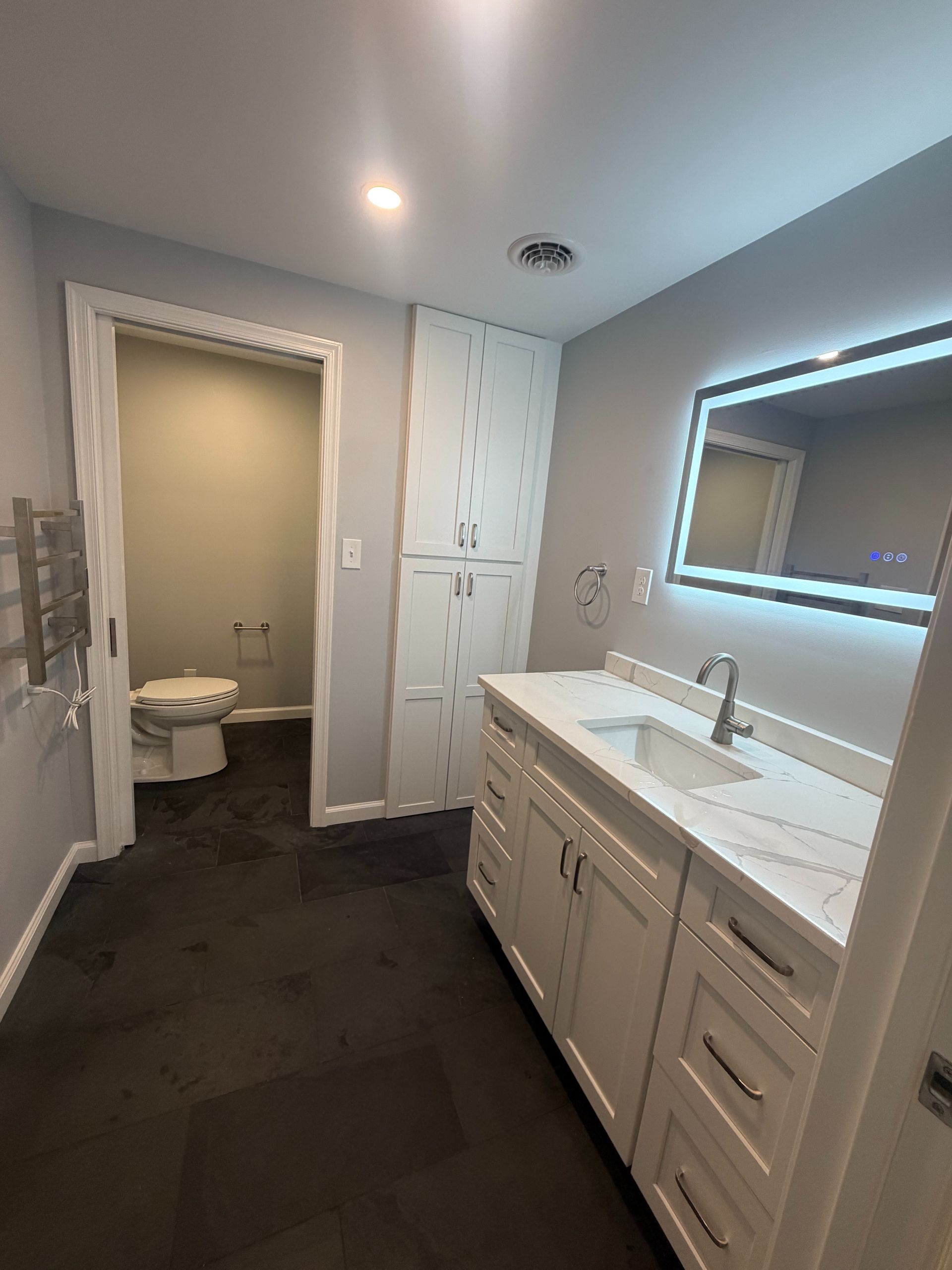 Modern bathroom with double vanity, illuminated mirror, and open toilet room in back