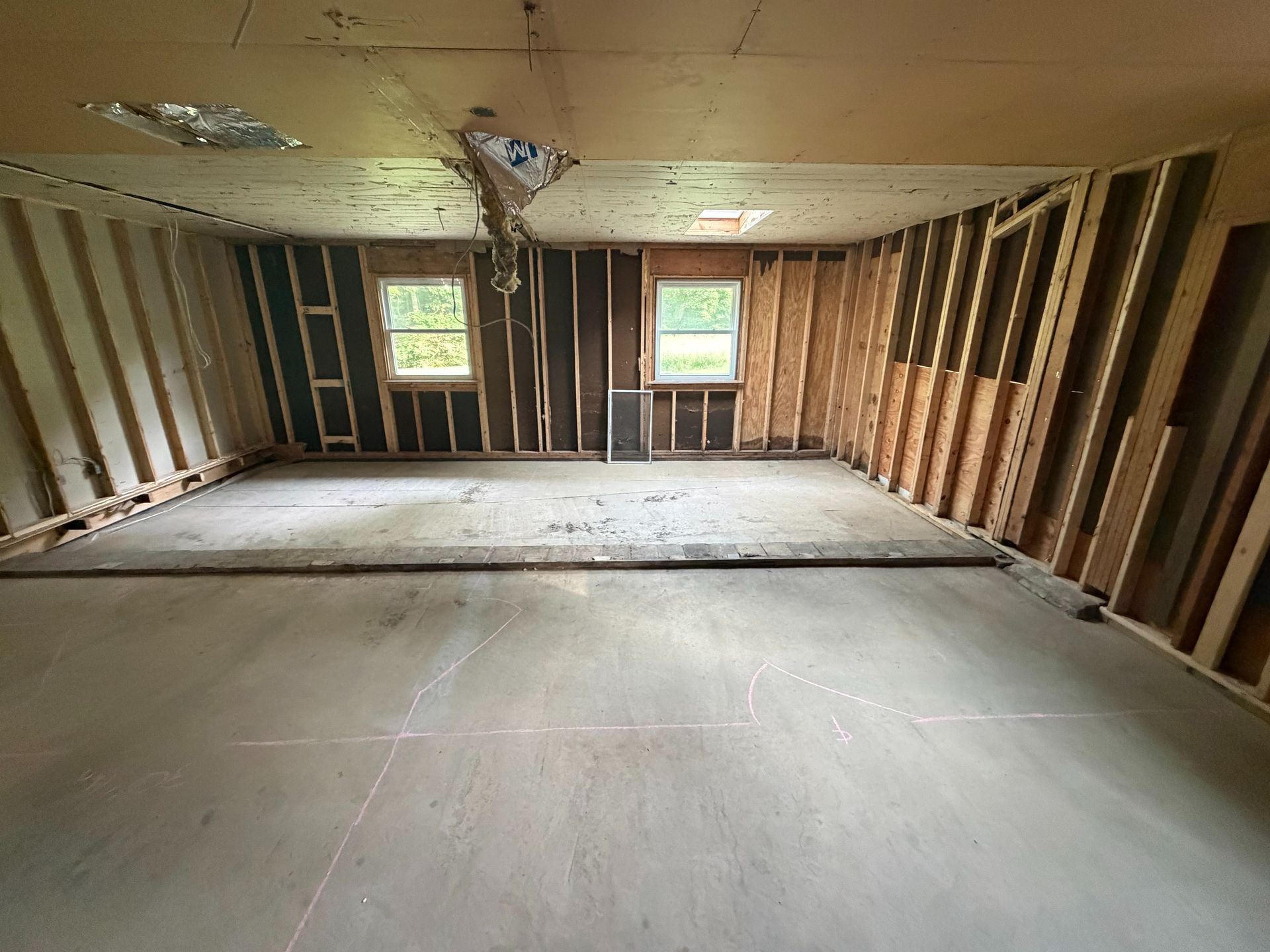 Empty unfinished room with exposed wood framing, bare floor, and two windows on the back wall