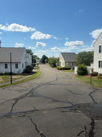 Quiet suburban street with white houses, cracked pavement, and a bright blue sky with scattered clouds