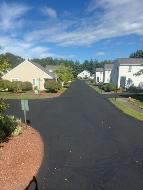Suburban street with freshly paved black road, white houses, green lawns, and a blue sky with clouds