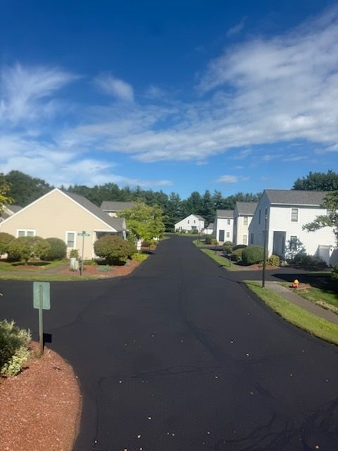 Suburban neighborhood street with white houses, a dark paved road, and blue sky with clouds