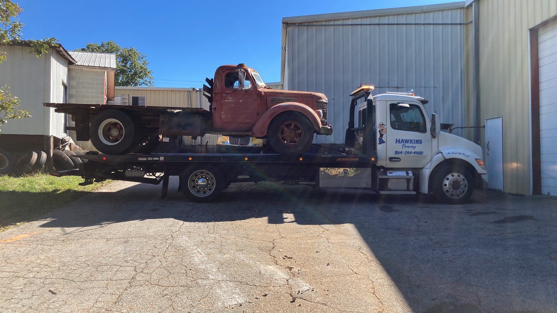 Tow truck carrying a rusty red pickup beside a metal building in a gravel lot.