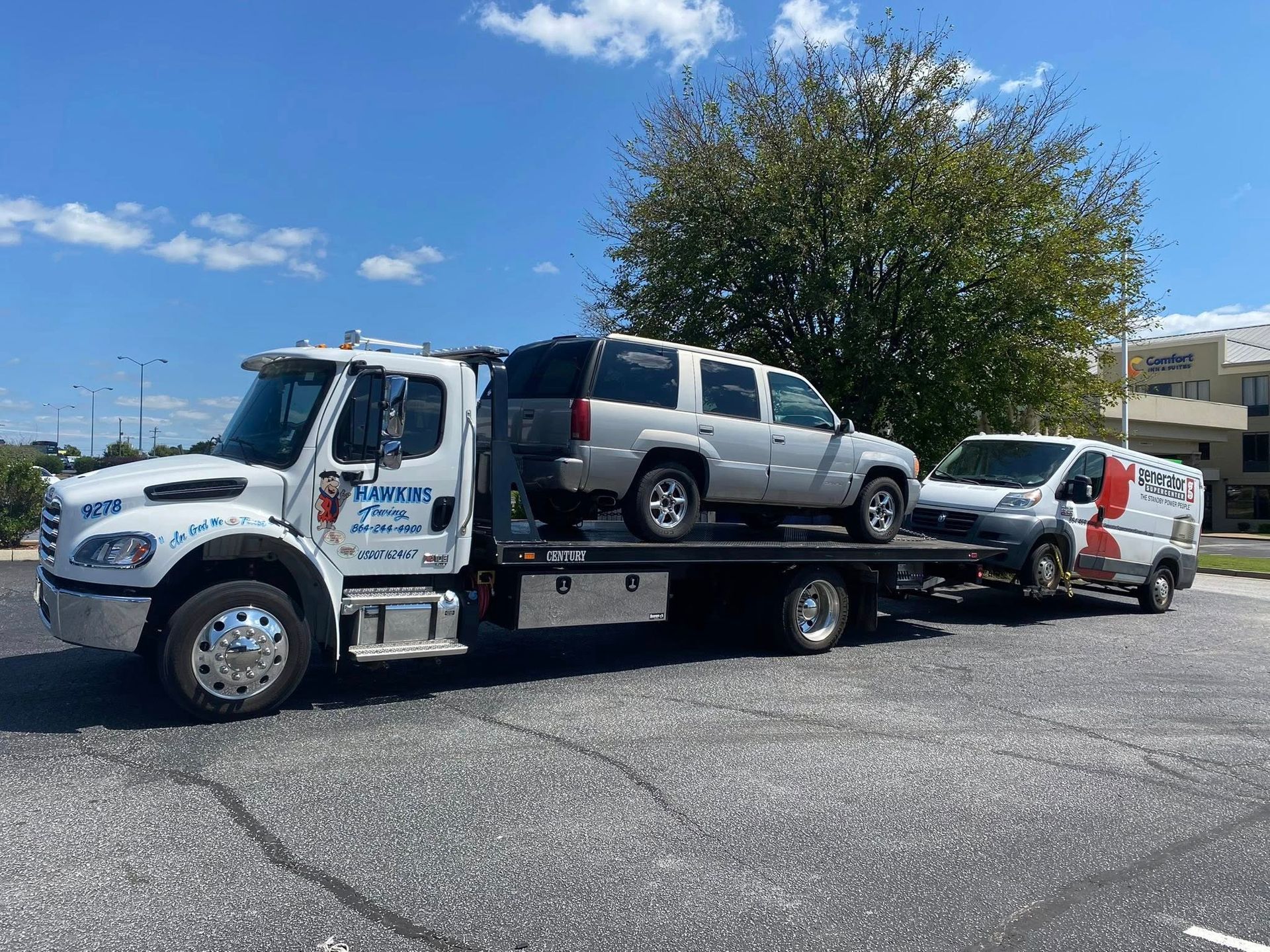 Tow truck carrying a silver SUV and white van in a parking lot on a sunny day