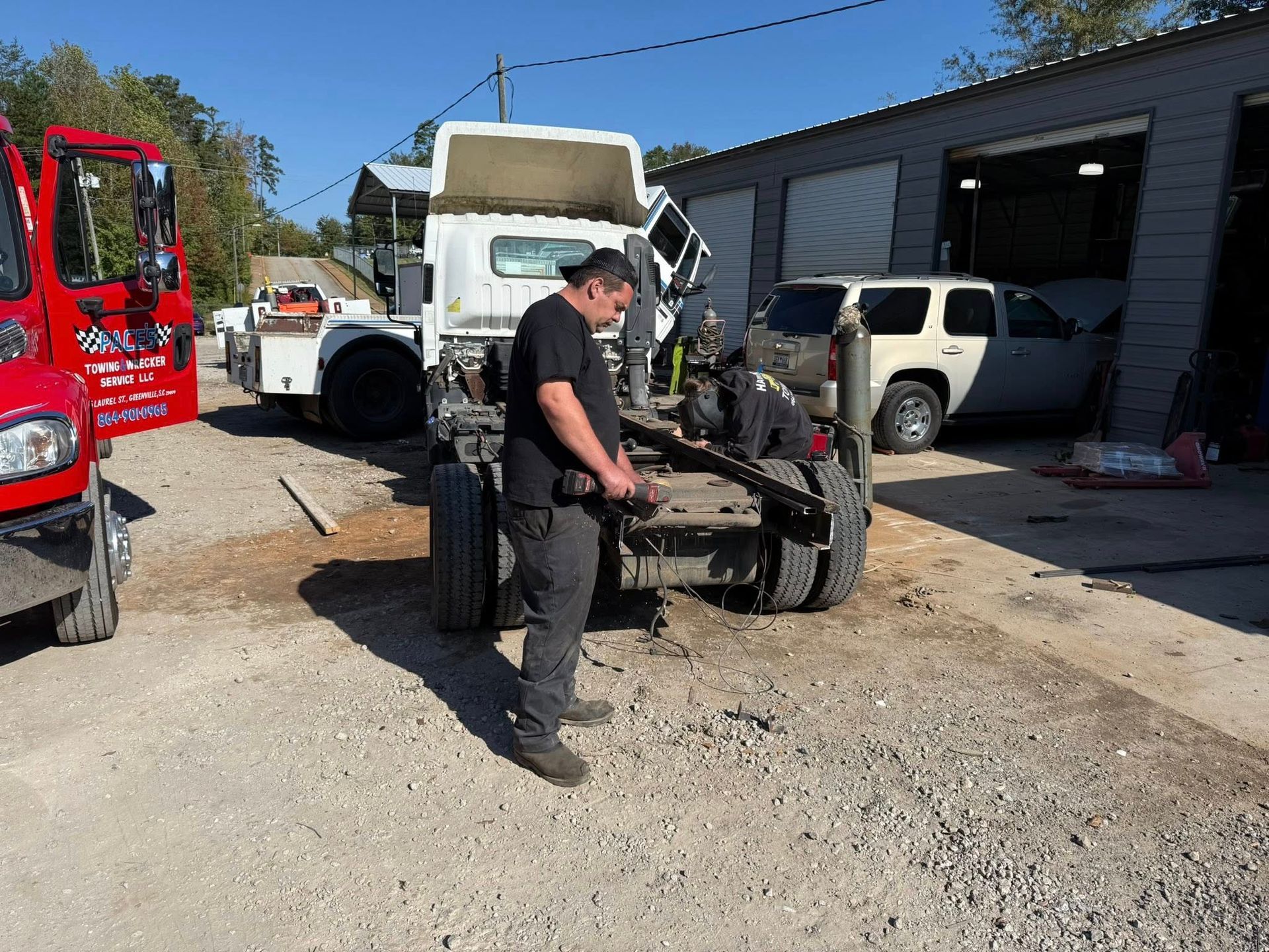 Mechanic in black shirt works on a vehicle chassis in a gravel auto yard with trucks and a garage nearby
