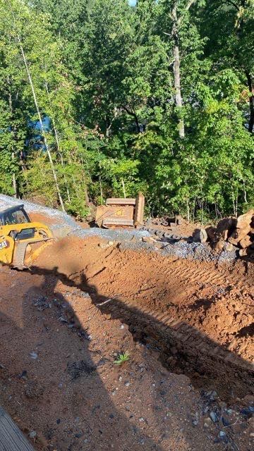 Excavator digging a dirt trench beside a wooded area on a sunny day