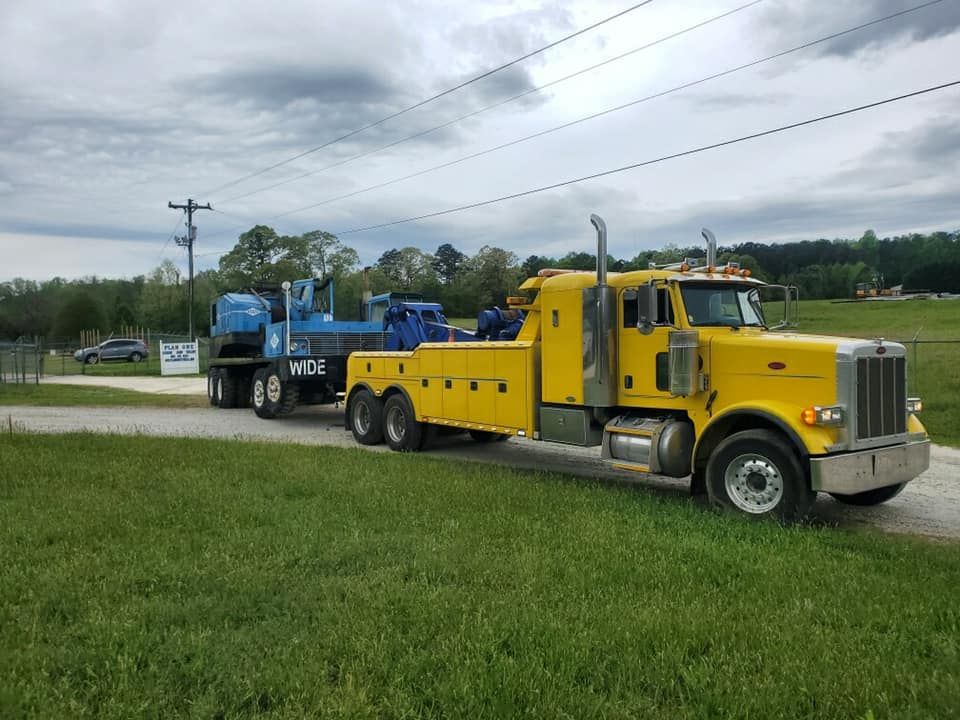 Yellow semi-truck hauling a blue industrial machine on a flatbed trailer along a grassy roadside