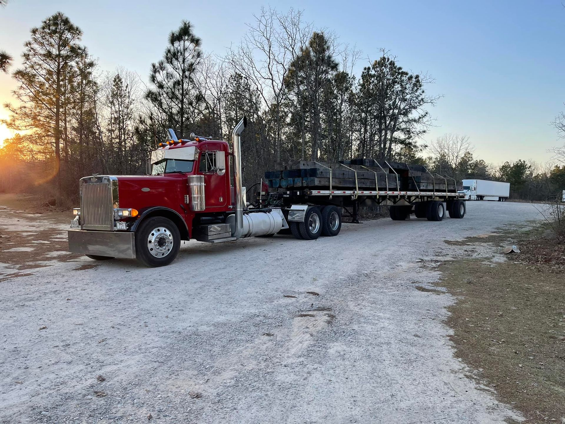 Red semi truck hauling a flatbed trailer on a snowy rural road at sunset