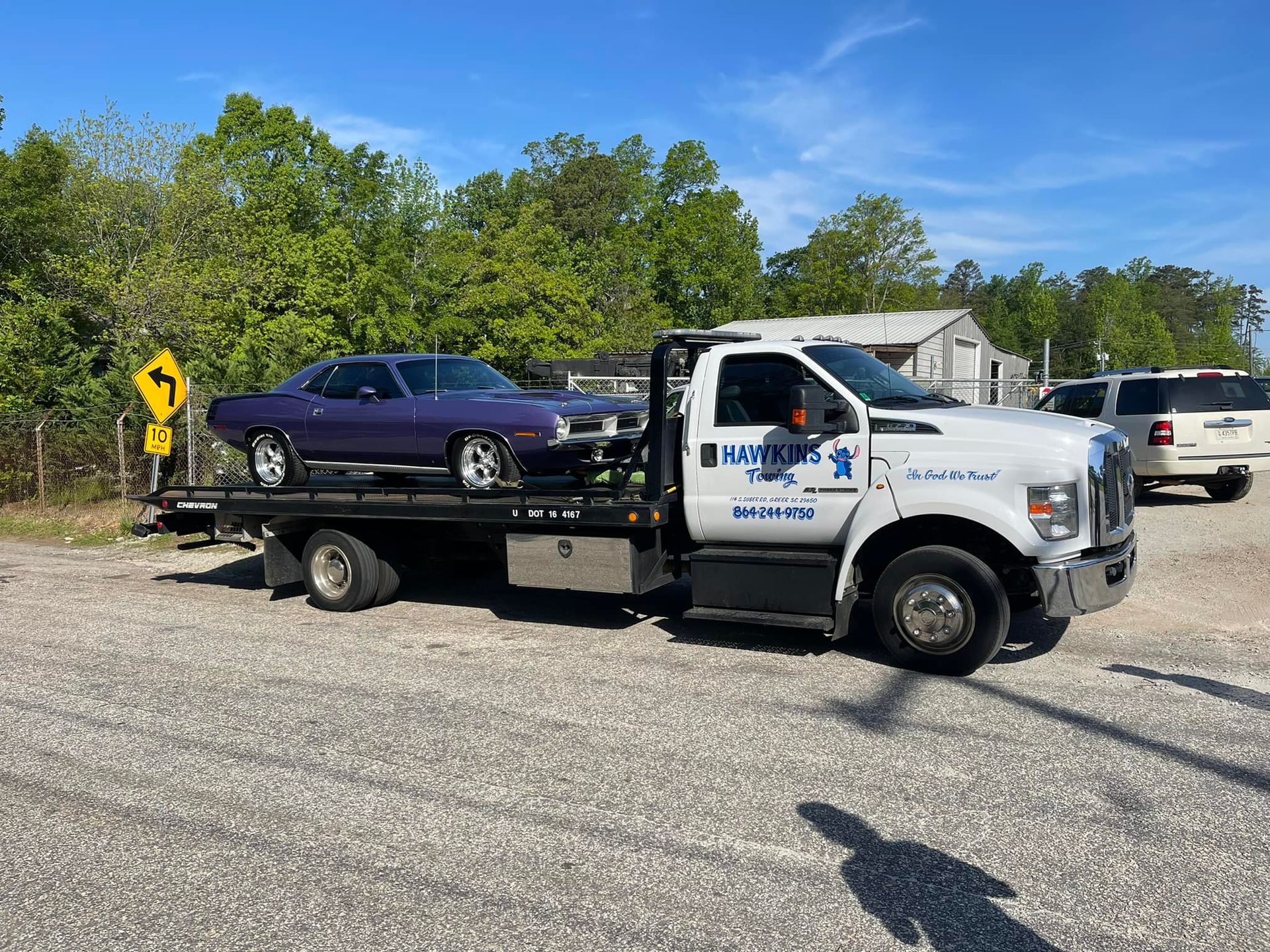 White tow truck hauling a purple car on a flatbed beside a roadside on a sunny day