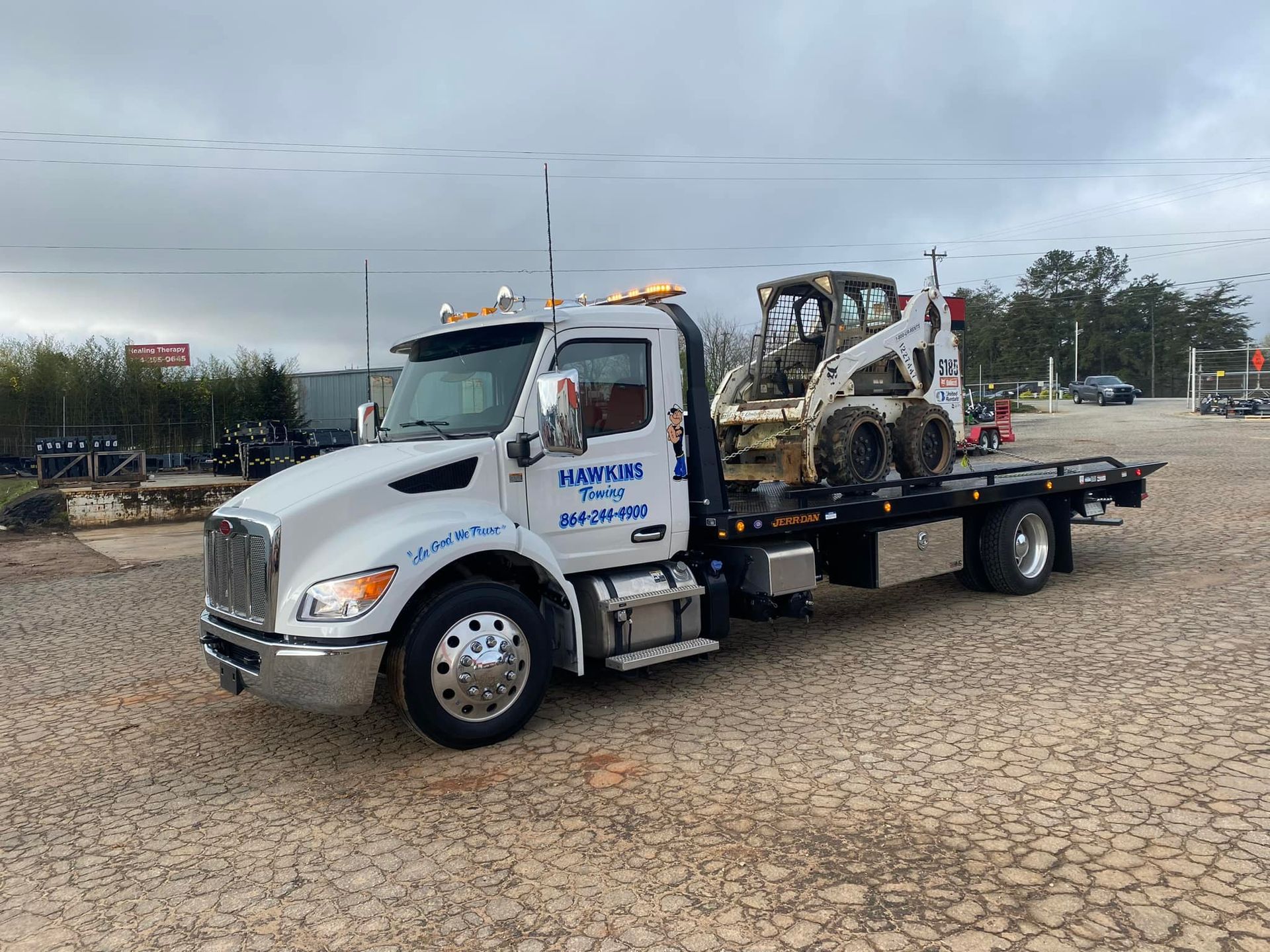 White flatbed tow truck with a wrecked car on a dirt lot under cloudy skies