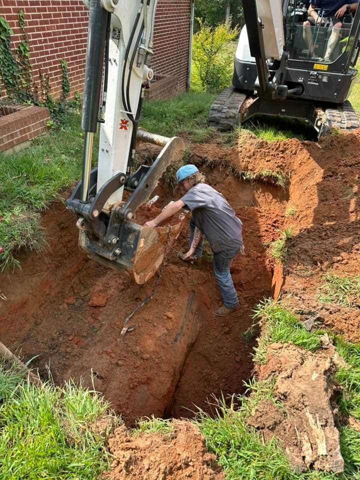 Excavator and worker digging a deep trench beside a brick building in muddy soil.