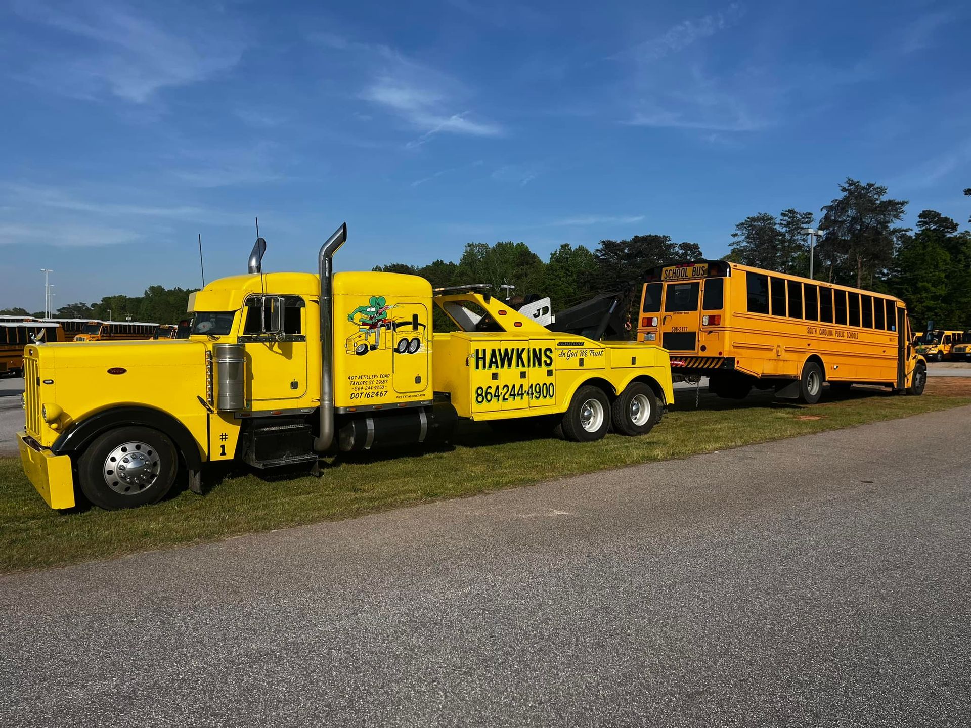 Yellow tow truck hauling a yellow school bus on a gravel lot under a blue sky