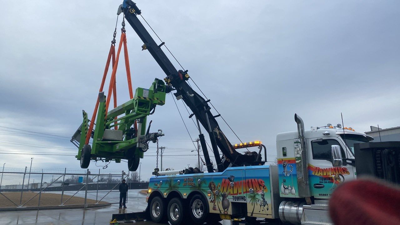 Large crane hoists a green amusement ride onto a decorated truck under a cloudy sky.