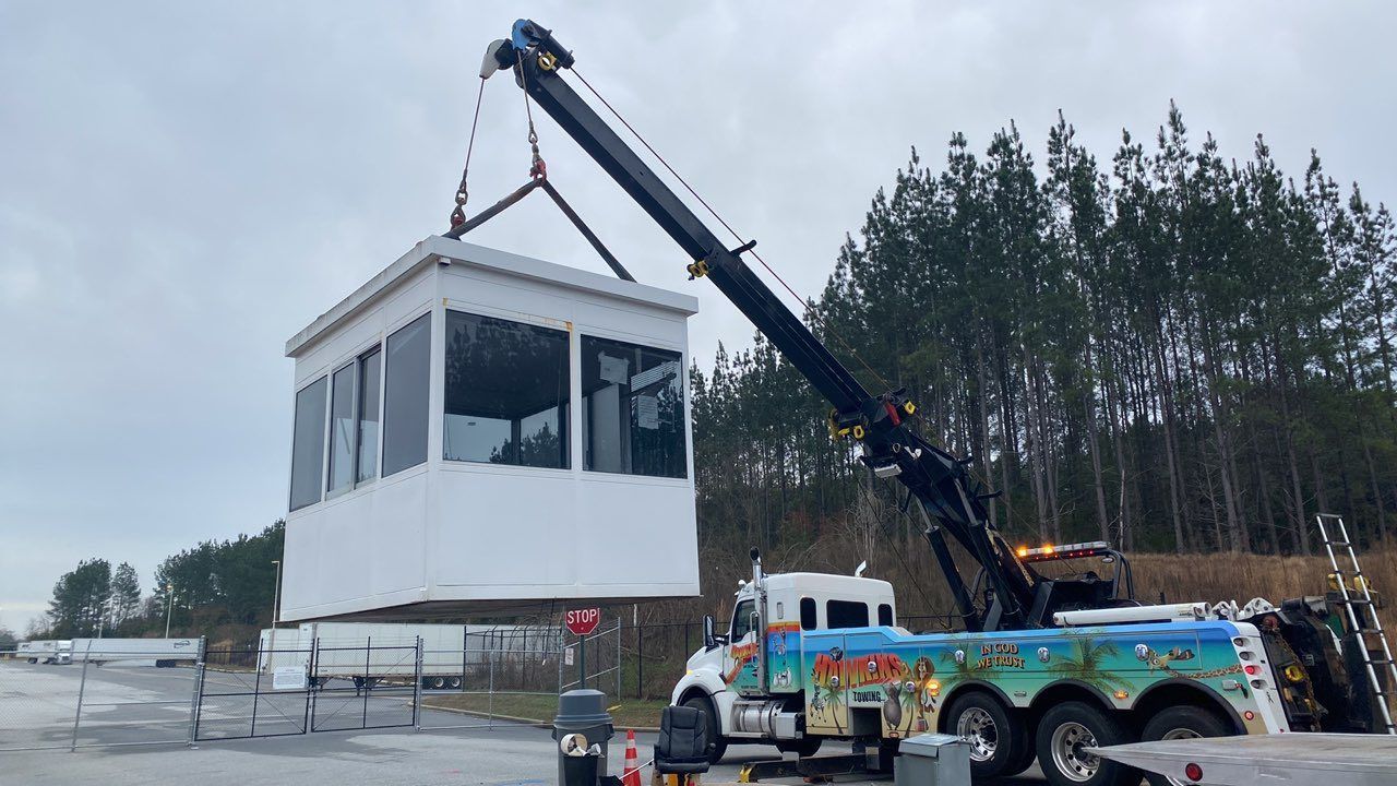 Crane lifting a white modular building beside a colorful truck on an overcast site